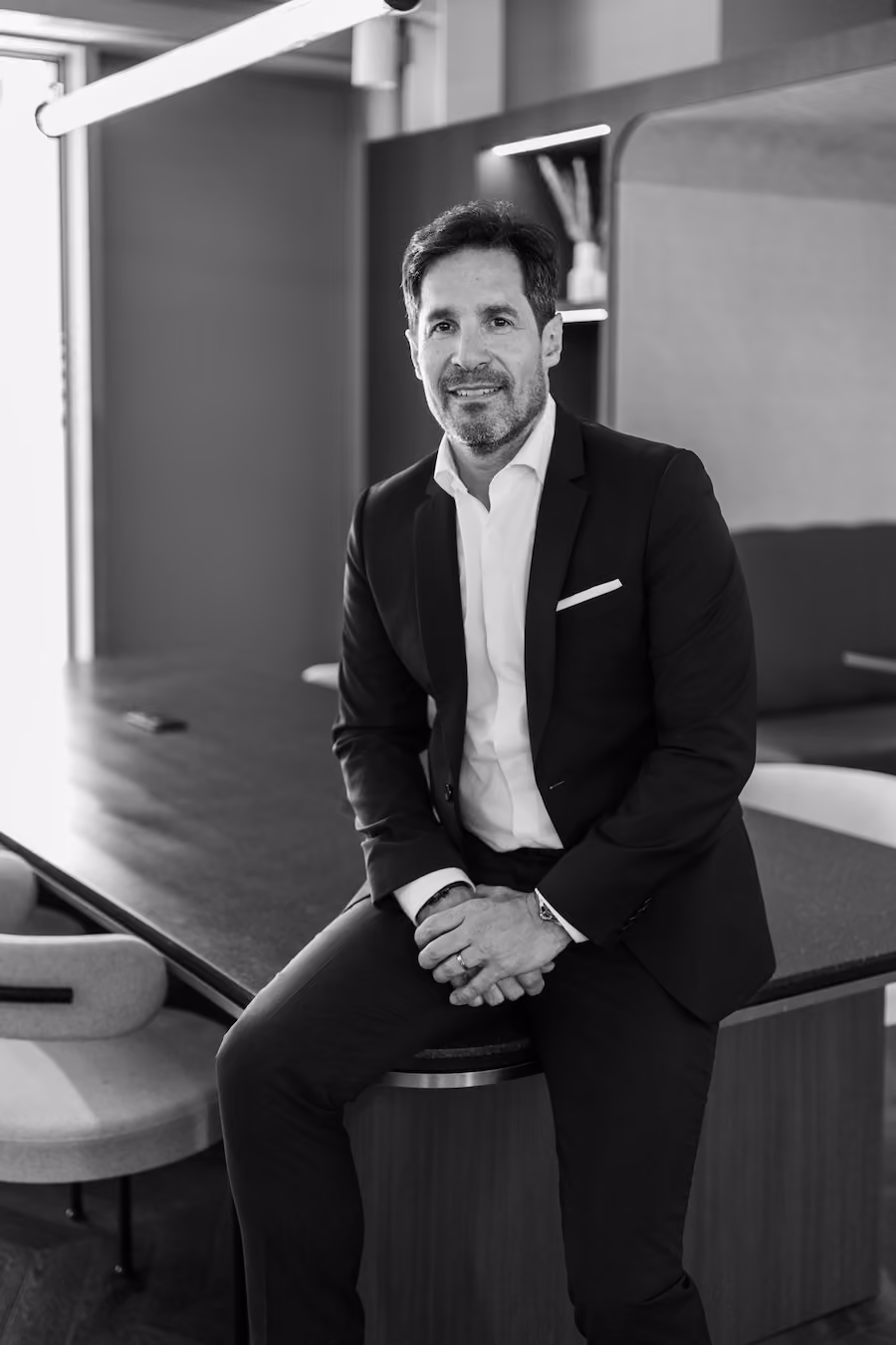Homme en costume assis sur un comptoir dans un bureau moderne, souriant vers la caméra.