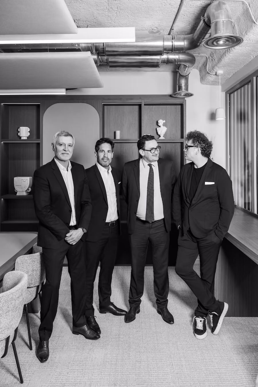 Quatre hommes en costumes debout dans un bureau moderne avec étagères et ventilation apparente.