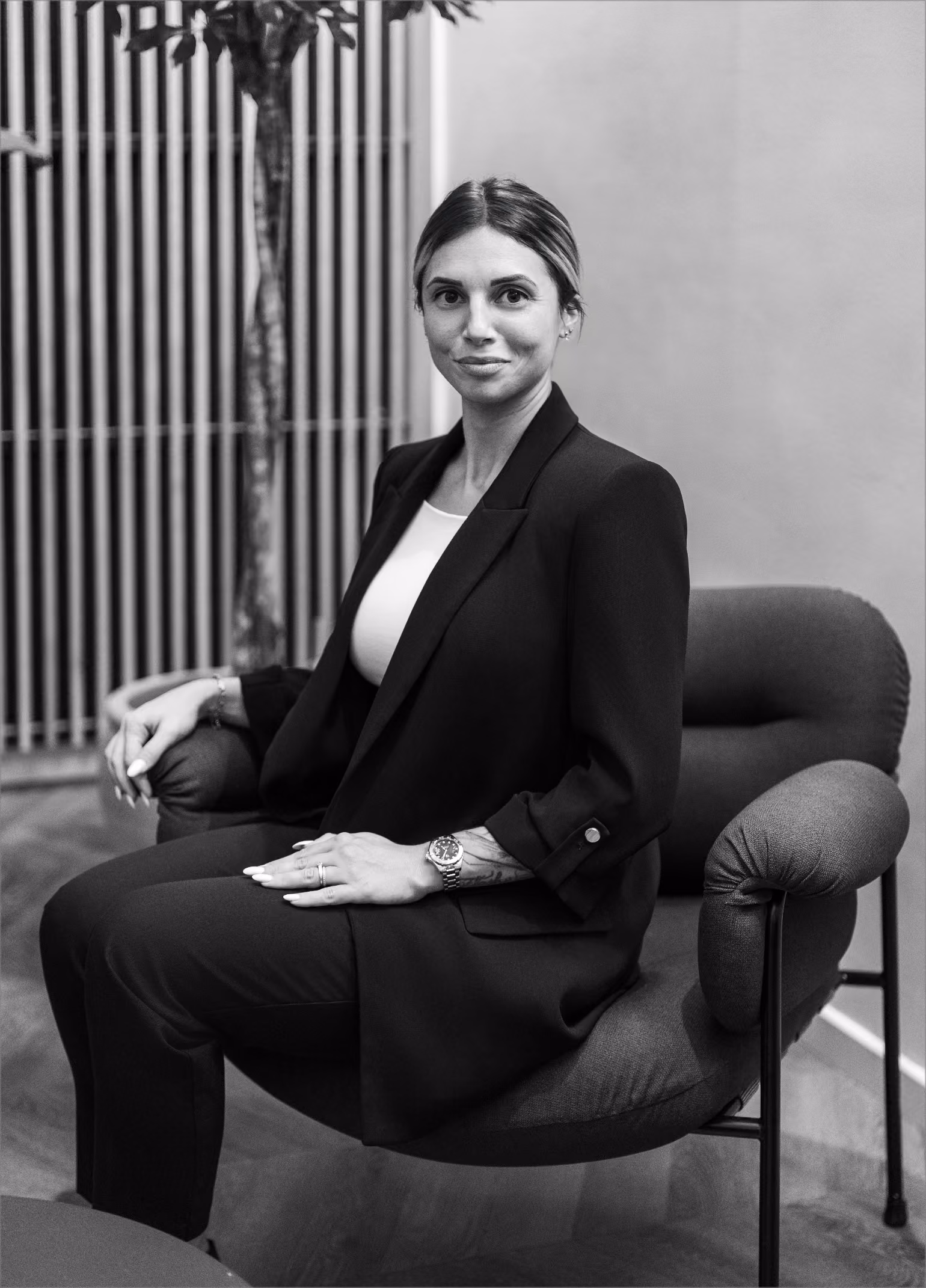 Femme assise sur une chaise moelleuse, portant un tailleur noir avec montre et tatouage visible.