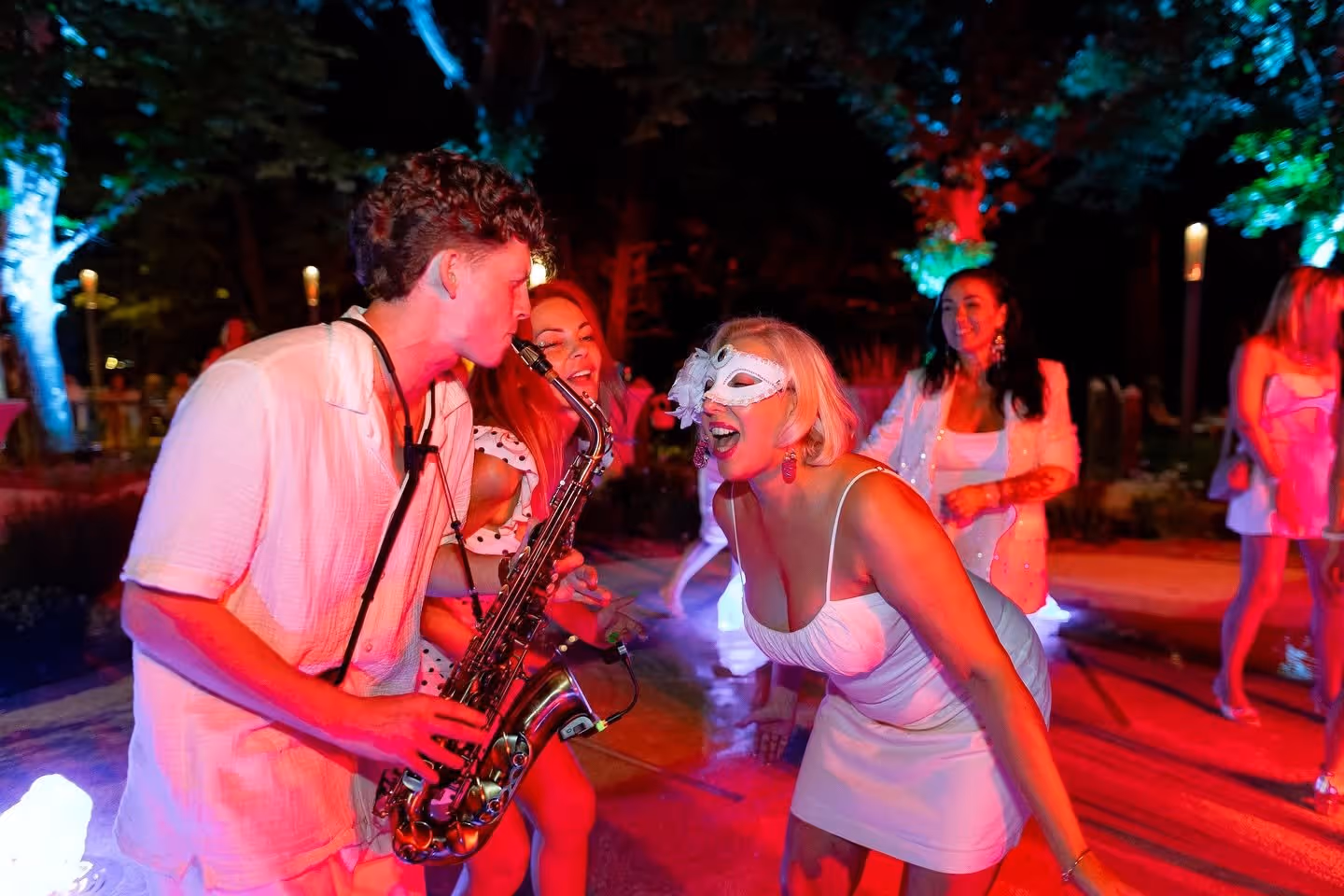 Man playing saxophone to a woman in a white dress and mask at a lively outdoor night party.