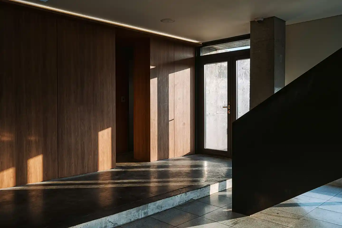 Sunlight casts shadows on a modern interior with wooden walls, a glass door, and dark staircase.