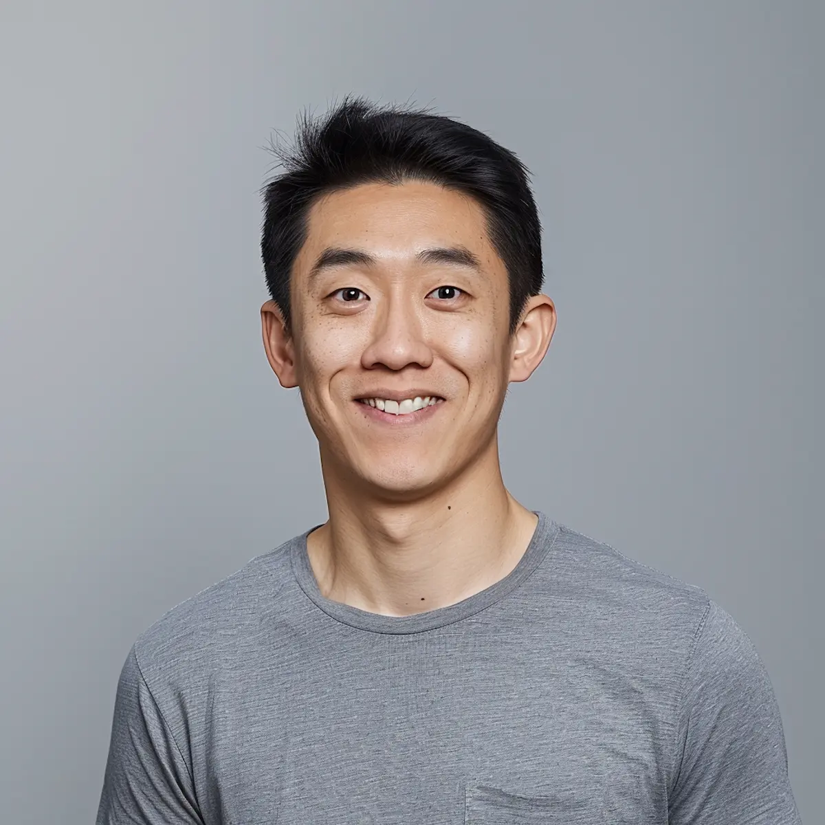 Young man with short black hair smiling in a gray T-shirt against a plain gray background.