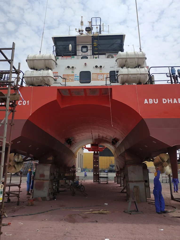 Large red ship in ICAD on dry dock with workers standing beneath its hull.