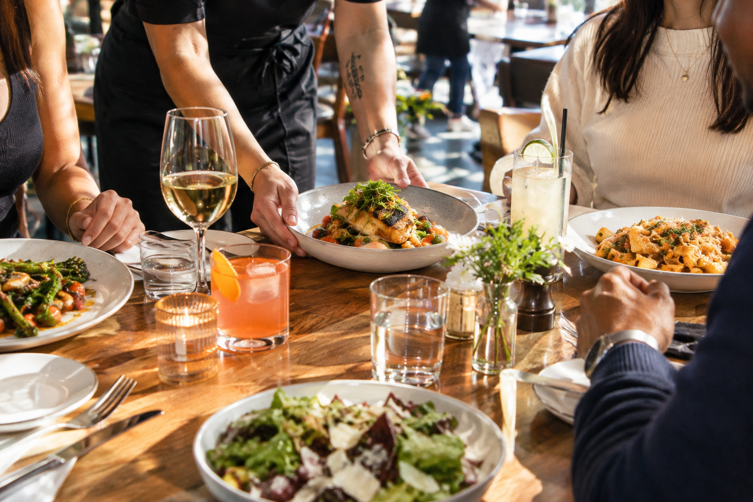 A server setting down plates at a busy bistro table with wine and cocktails