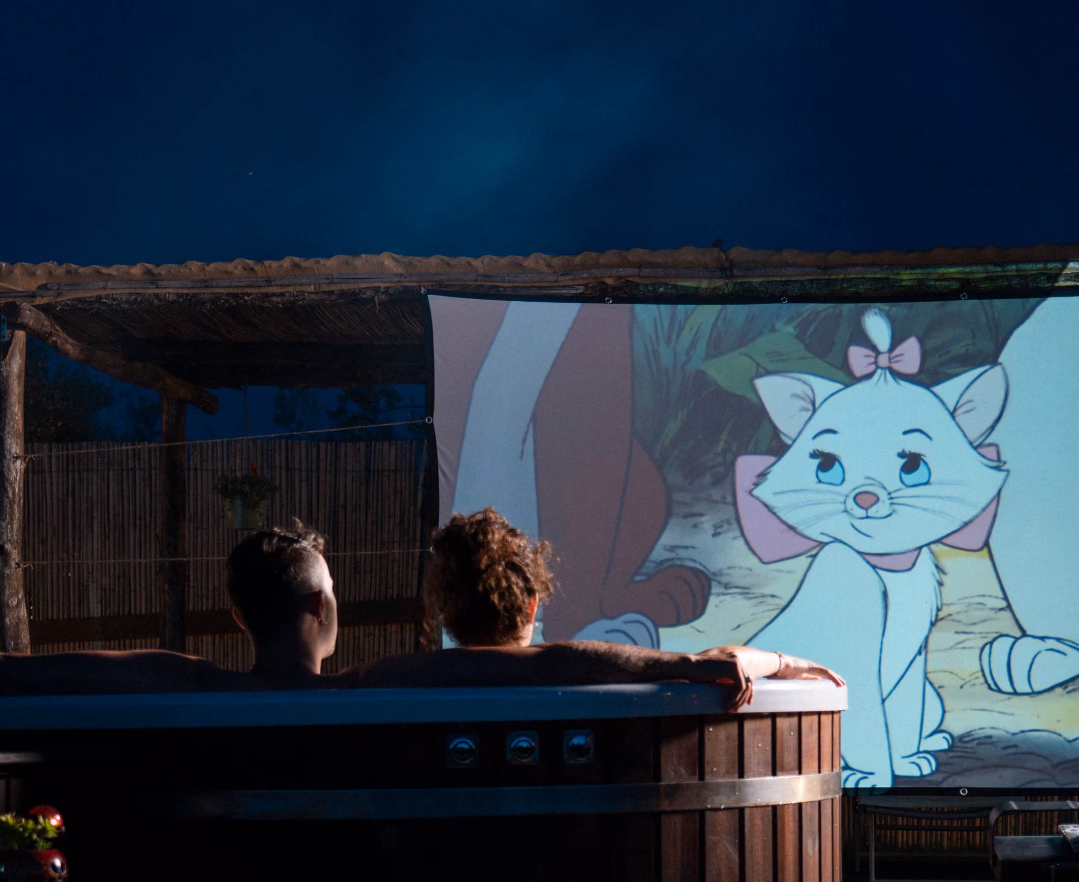 Una coppie immersa nell'hot tub (o jacuzzi o bagno caldo) fuori dalla propria yurta, mentre guarda un film al cinema privato al Glamping per coppie Due cuori e una Yurta