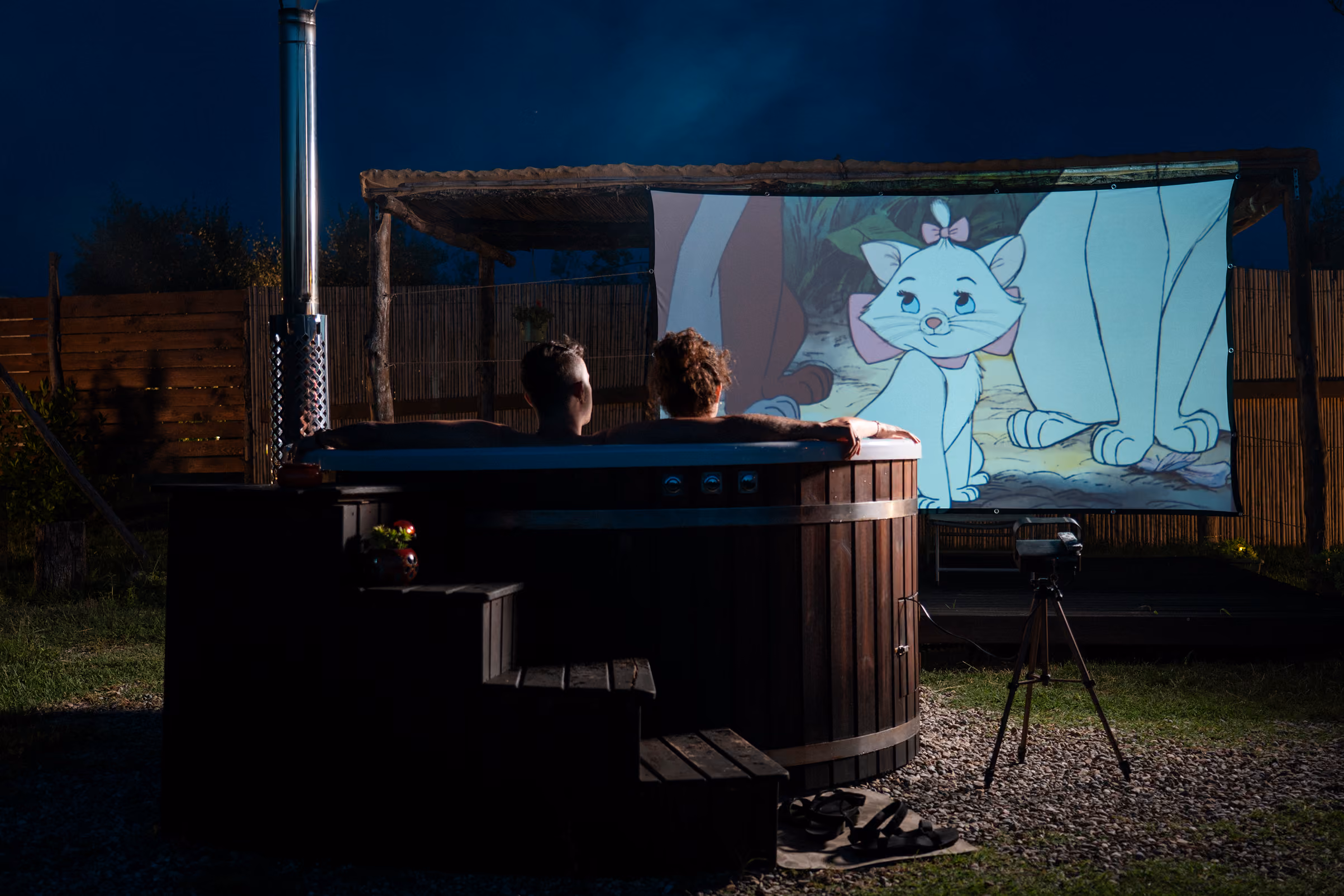 Una coppia dentro un'hot tub (jacuzzi o bagno caldo) guarda un film al cinema privato fiori dalla propria yurta al Glamping per coppie Due cuori e una Yurta