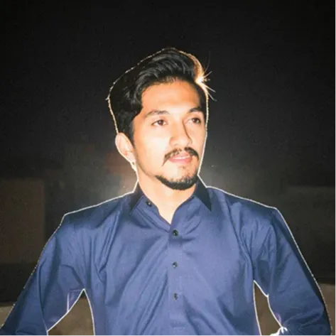 Young man with neatly styled hair and a trimmed beard wearing a blue button-up shirt at night.