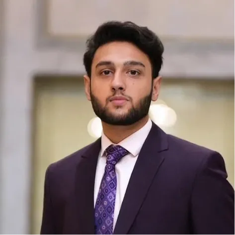 Young man with dark hair and beard wearing a purple suit and patterned tie.