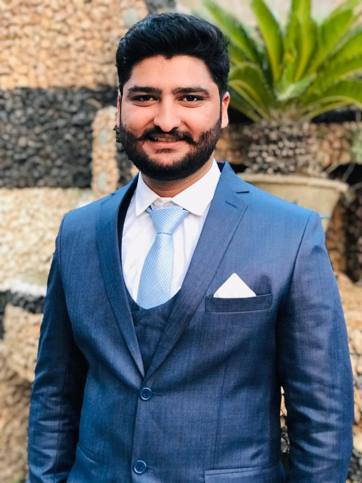 Smiling man with beard wearing a blue suit, light blue tie, and white shirt outdoors.