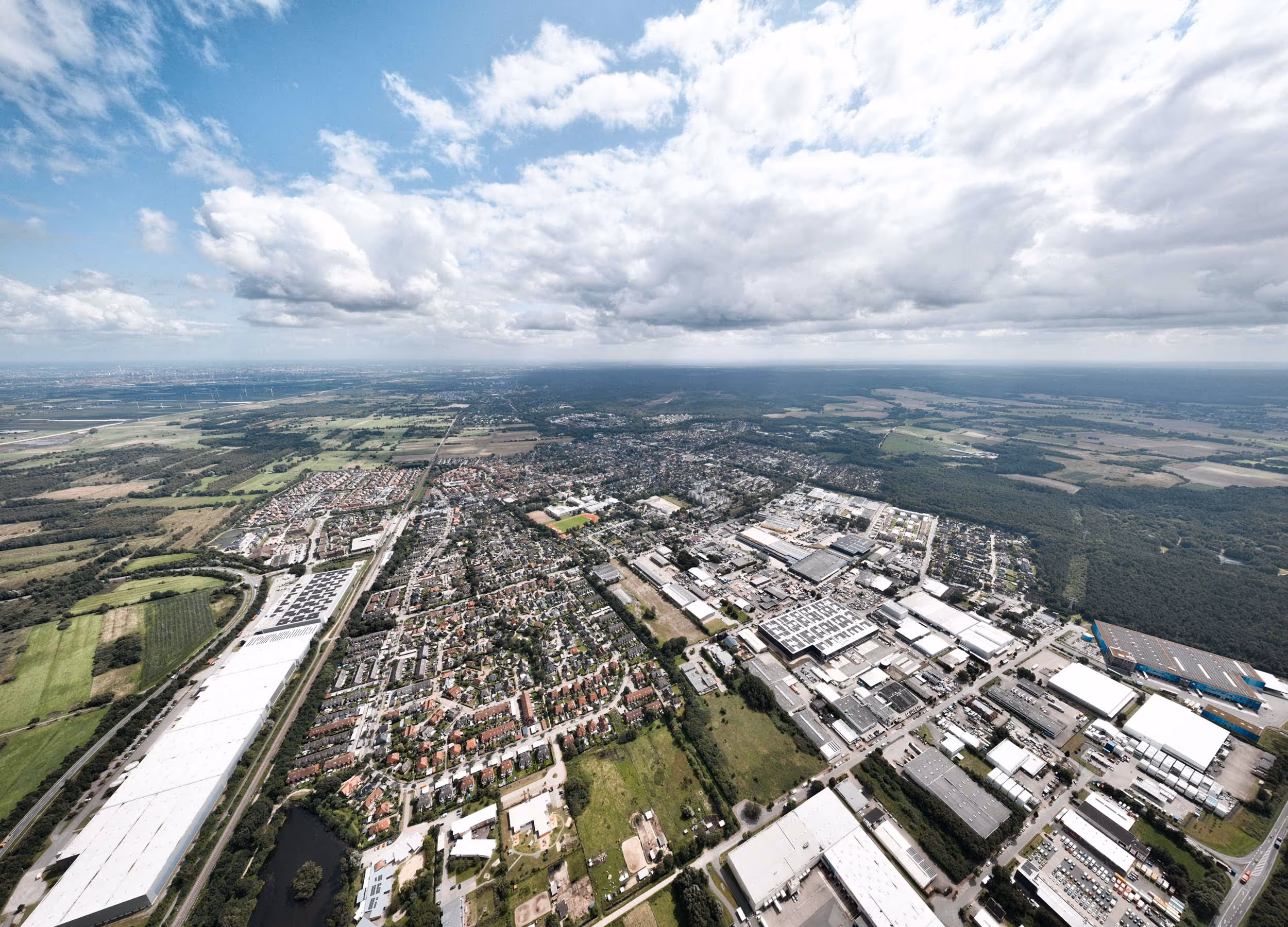 Luftaufnahme einer Stadt mit einer Mischung aus Wohngebieten, Industrieanlagen und umliegenden Feldern unter einem teils bewölkten Himmel.