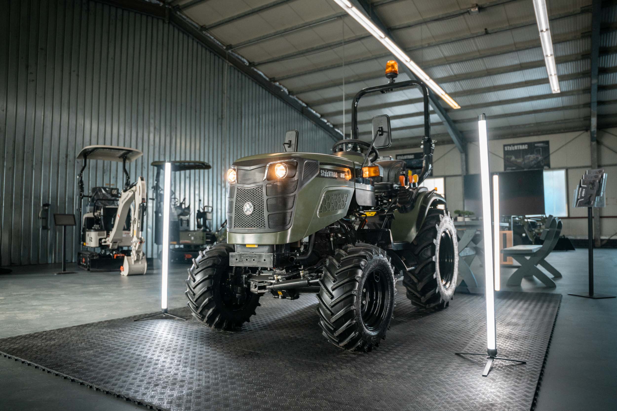 Grüner StartTrac-Traktor mit eingeschalteten Scheinwerfern in einer gut beleuchteten Halle.