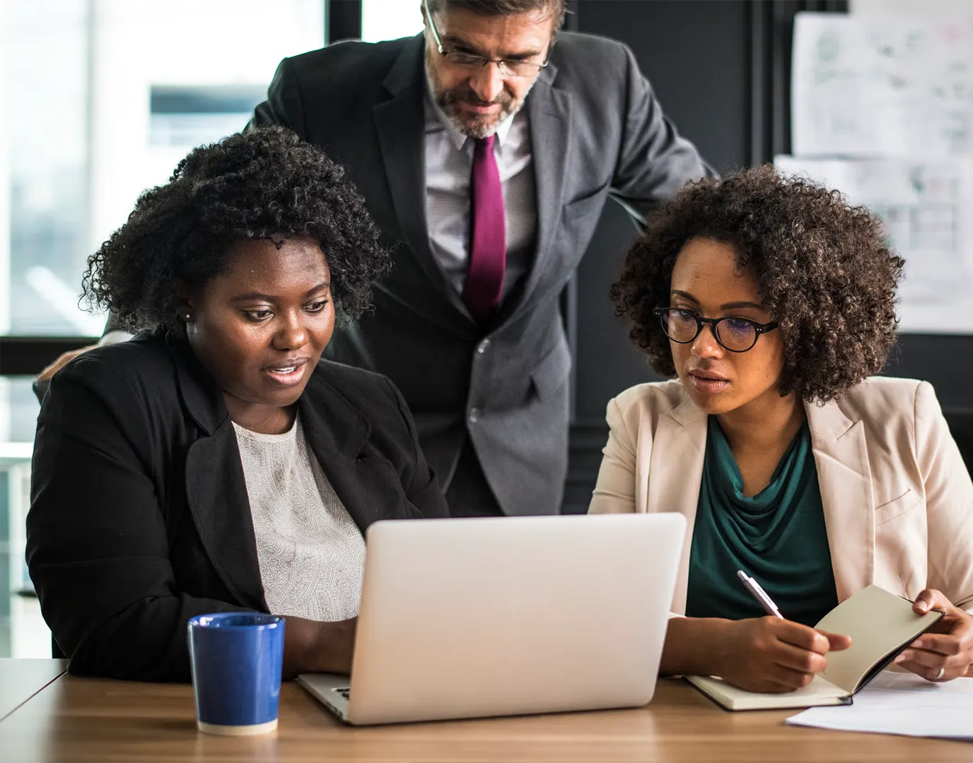 Two women and a man working together at a laptop in an office setting, one woman taking notes.