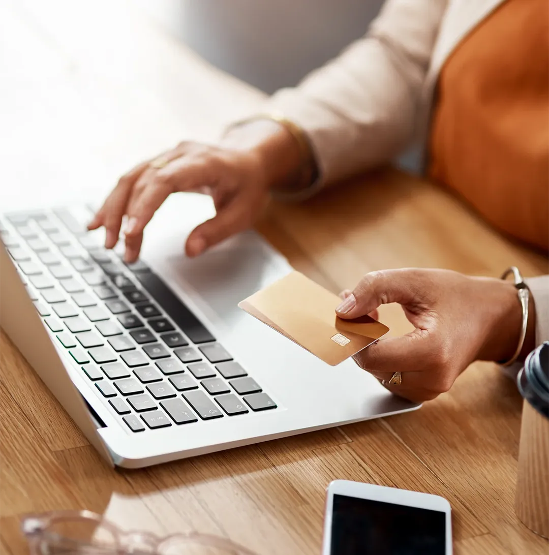 Person holding a credit card and typing on a laptop keyboard on a wooden table with a phone nearby.