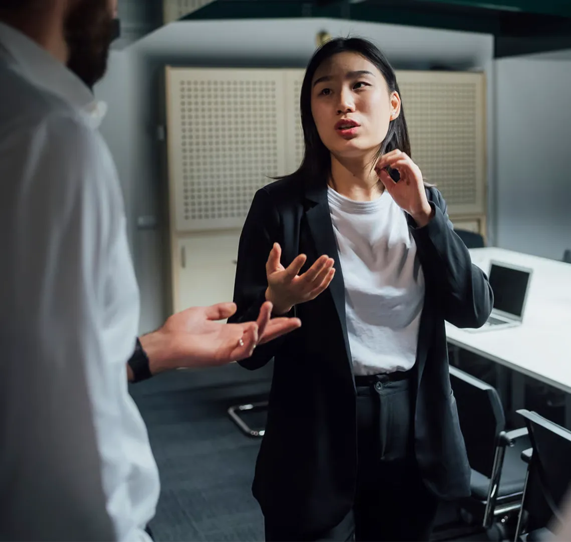 Two colleagues in business attire having a serious conversation in an office meeting room.