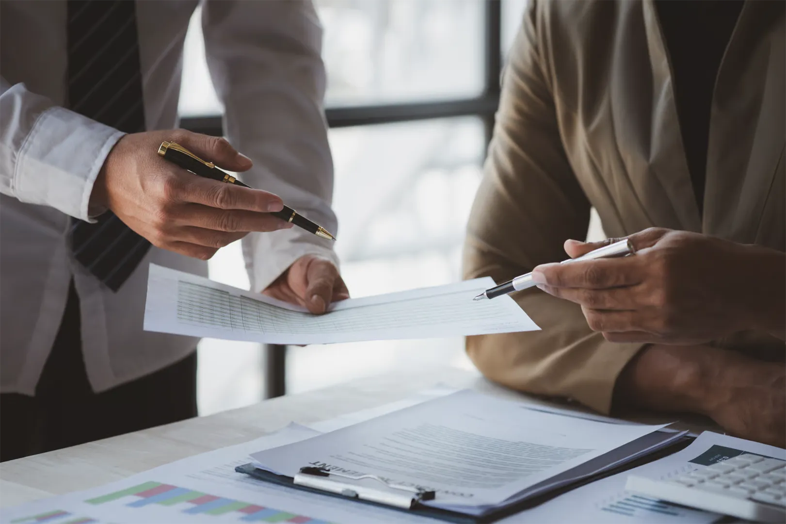 Two people in business attire reviewing and pointing at a document with pens in an office.