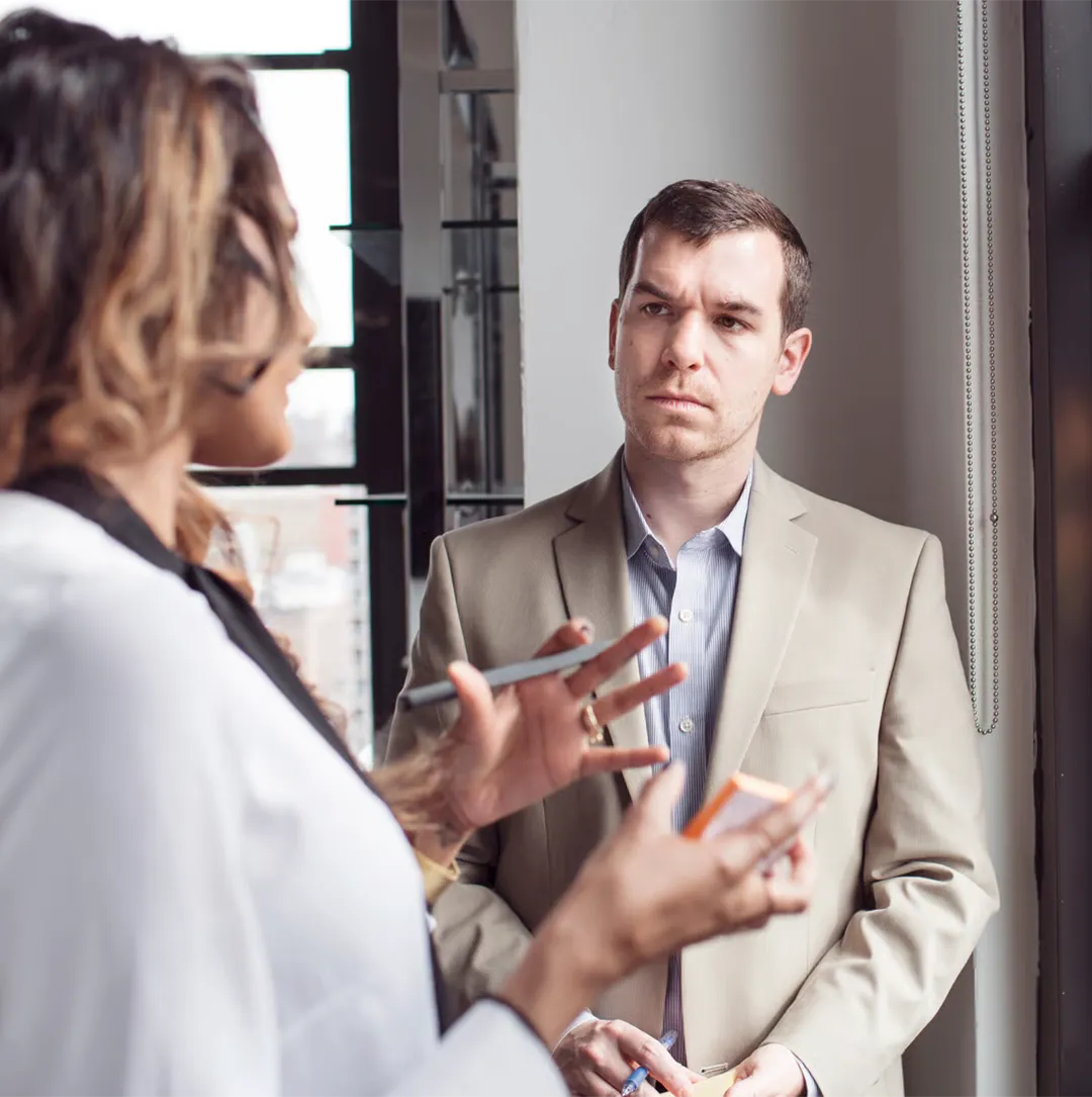 A woman talks and gestures while a man in a beige suit listens attentively near a window.