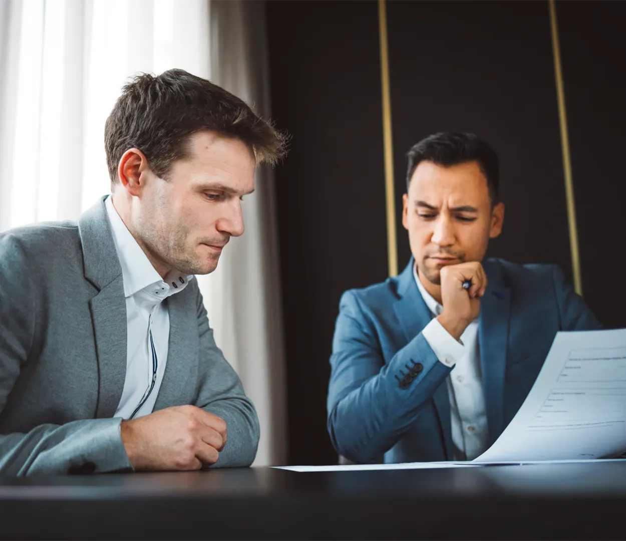 Two men in business suits reviewing and discussing documents at a table near a window.