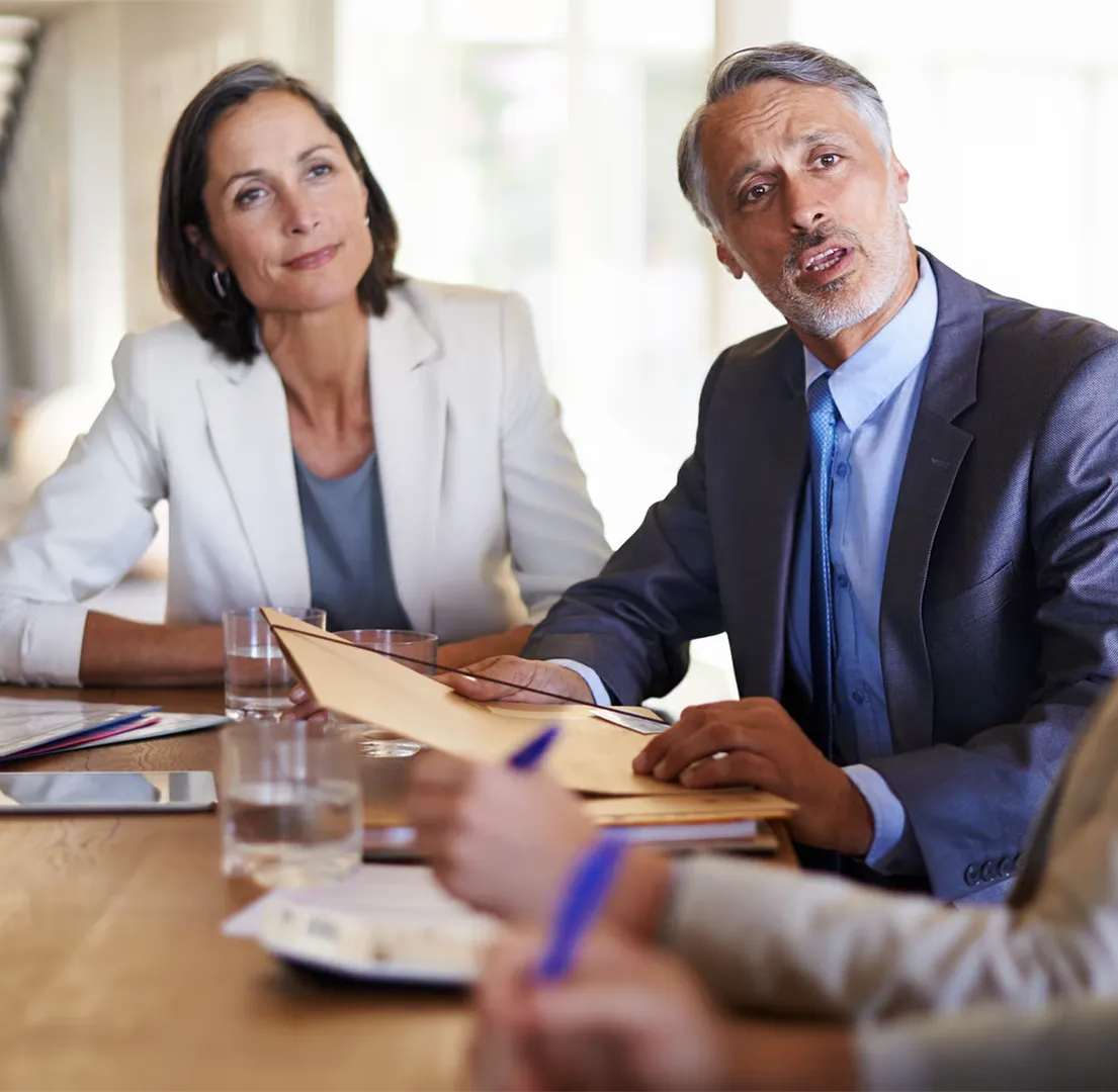 Man in suit holding folder speaking, woman in light blazer listening at business meeting table.