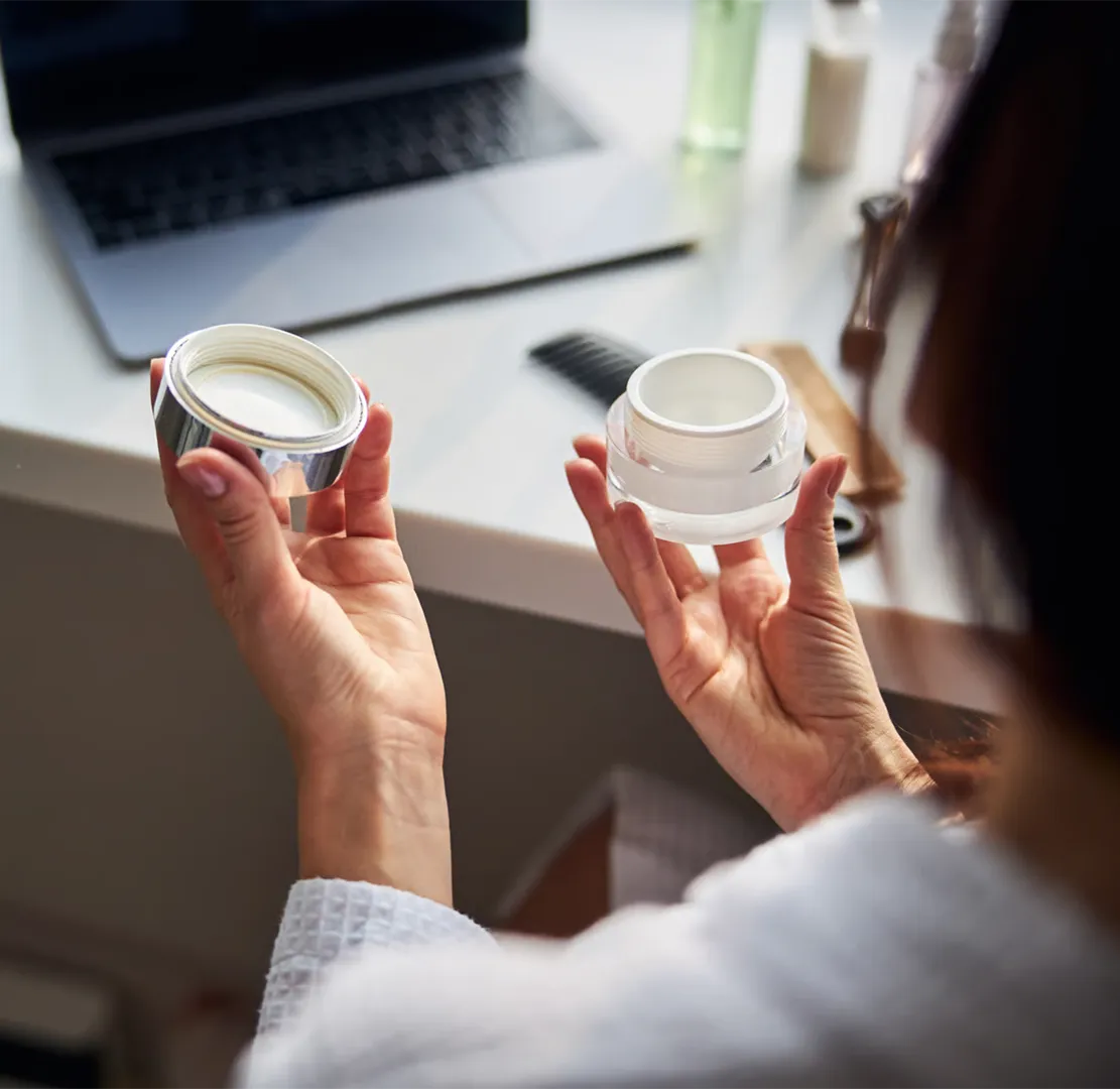 Person holding an open jar of white cream with a laptop and skincare items on a desk.
