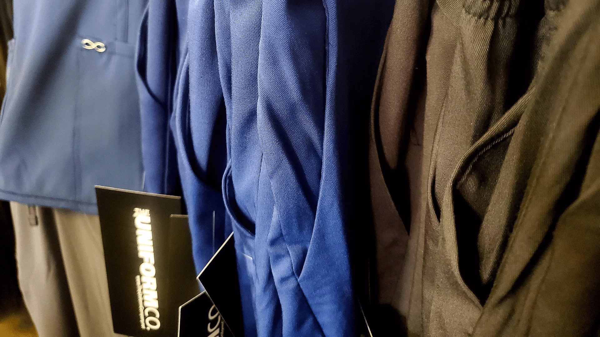 Close-up of blue and gray uniform jackets hanging with tags from The Uniform Co.