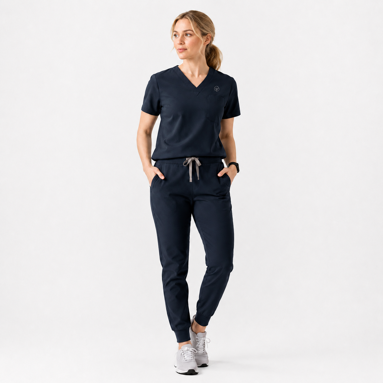 Woman in navy blue medical scrubs with hands in pockets, looking sideways, wearing white sneakers.