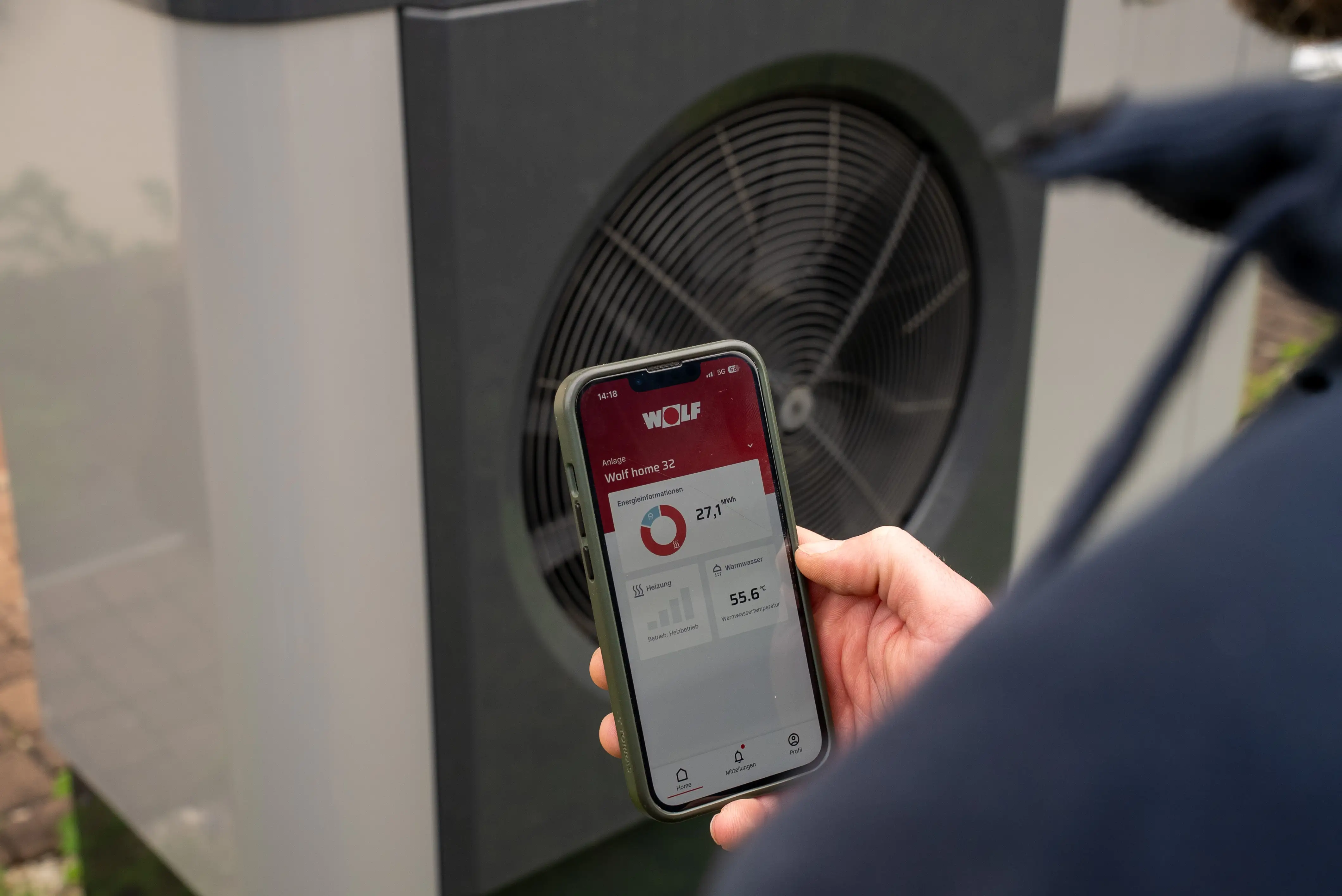 Hand hält Smartphone mit Wolf Wärmepumpen-App vor einem Gerät mit großem Lüfter.
