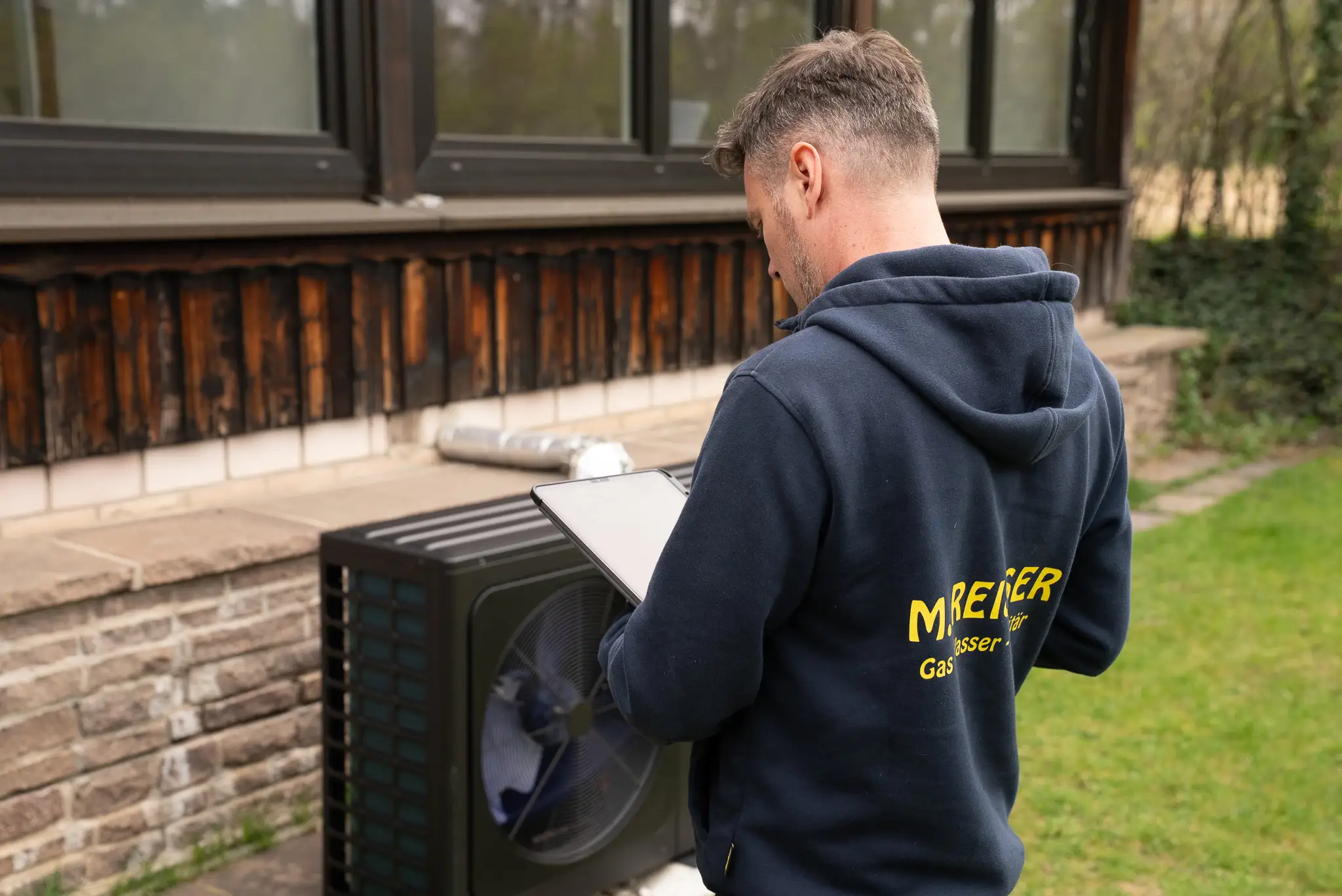 Ein Techniker mit Tablet kontrolliert eine Luftwärmepumpe vor einem Haus mit Holzverkleidung.