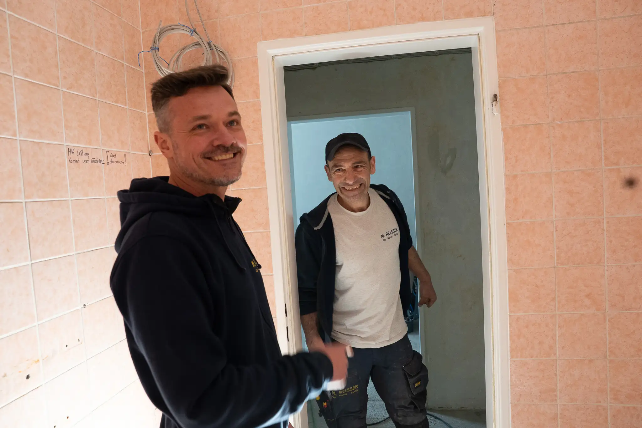 Zwei Handwerker schneiden ein Brett mit einer Kreissäge im Freien, einer hält Staubsaugerrohr.