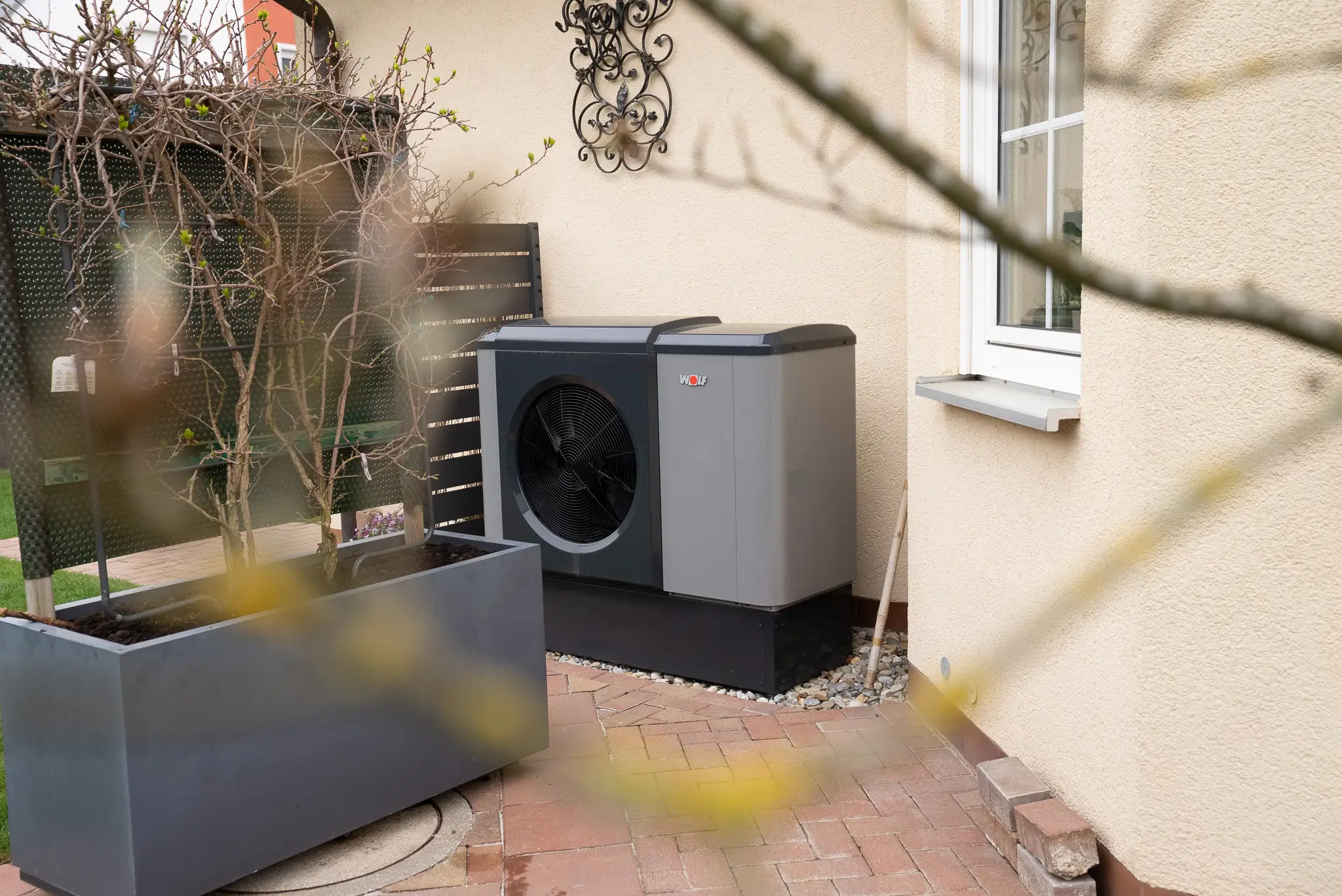 Moderne Wärmepumpe neben Hauswand auf gepflastertem Boden mit Blumenkasten im Vordergrund.