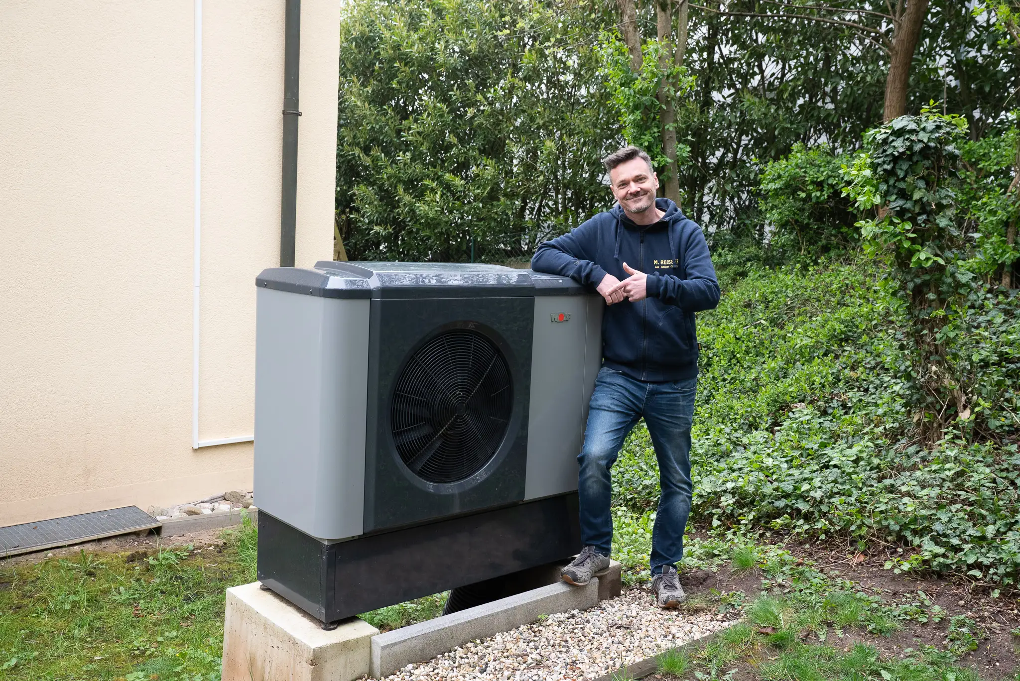 Mann lehnt lächelnd an grauer Wärmepumpe neben Haus, umgeben von Garten und Bäumen.