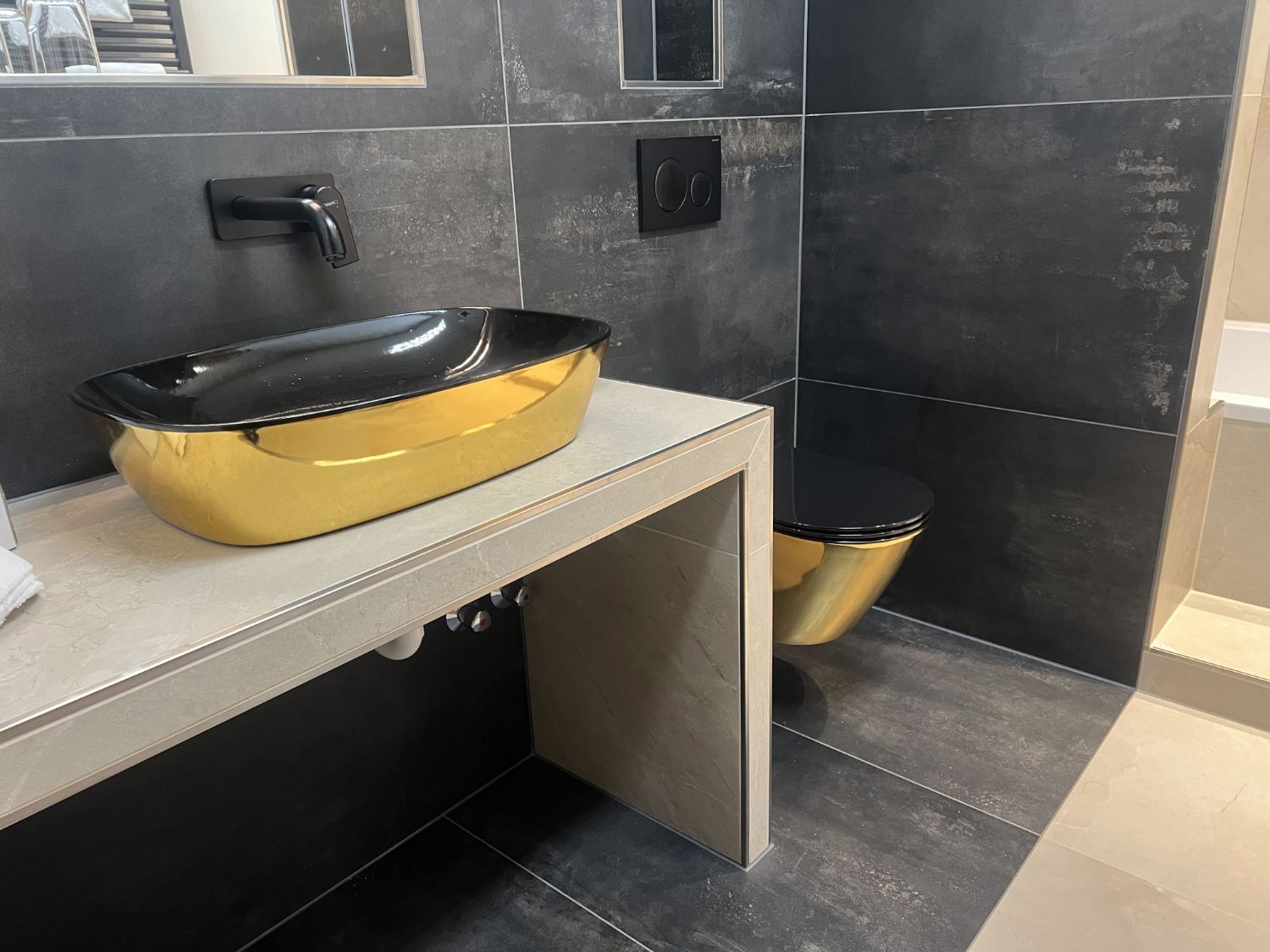Modernes Badezimmer mit goldfarbenem Waschbecken und Toilette, schwarzen Armaturen und dunklen Fliesen.