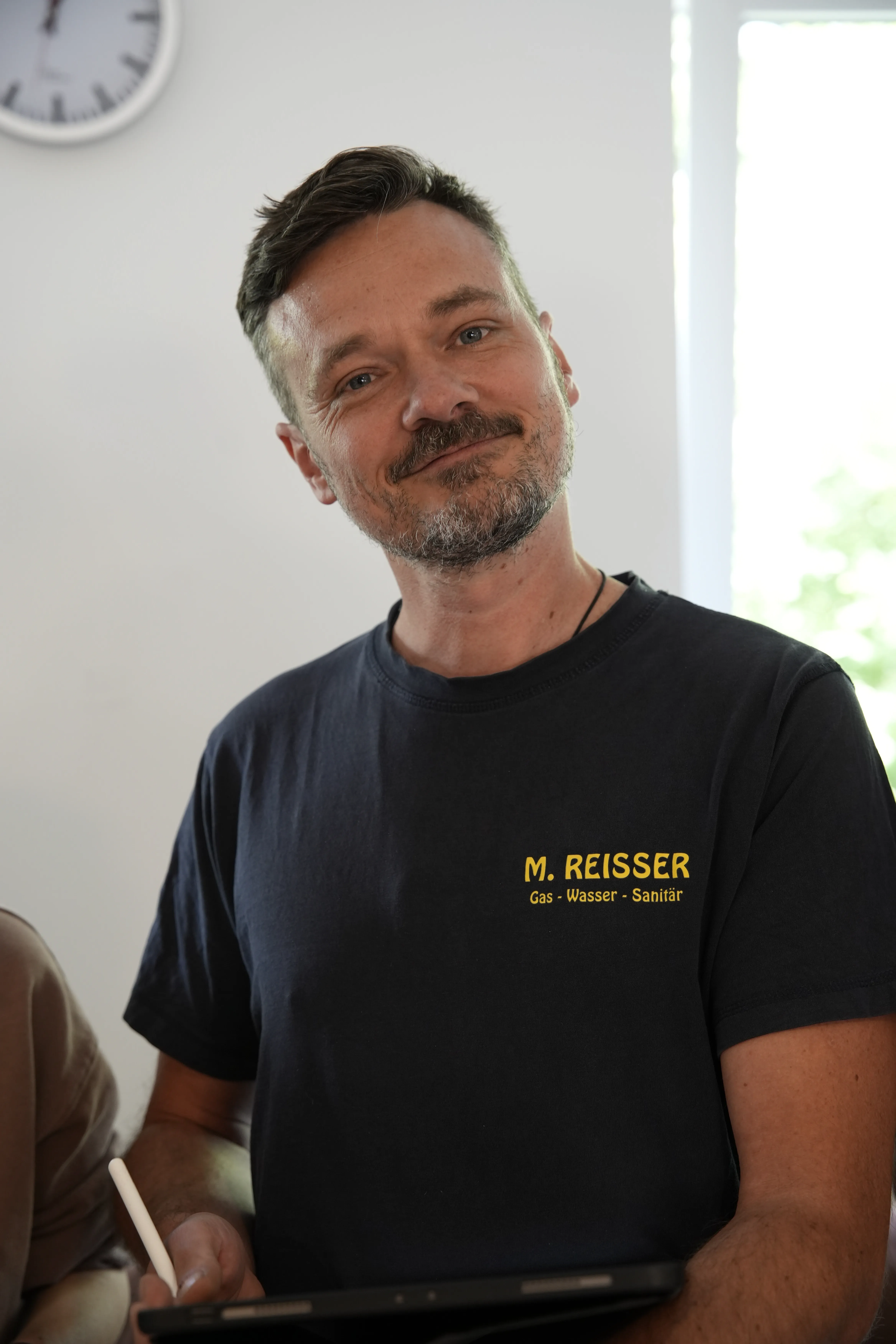 Mann mit dunklem T-Shirt mit Aufschrift M. Reisser Gas Wasser Sanitär, der ein Tablet mit Stift hält.