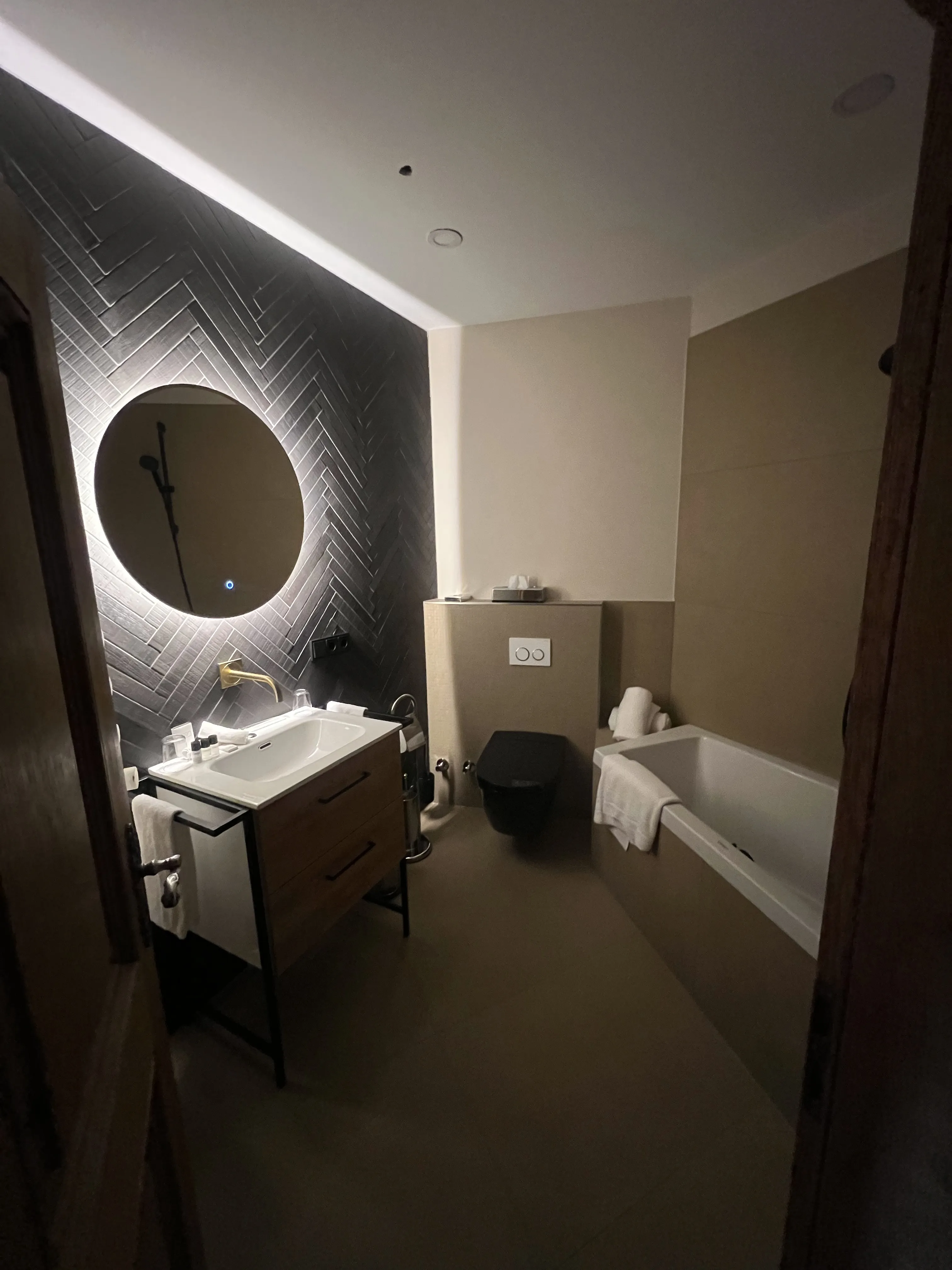 Modernes Badezimmer mit beleuchtetem runden Spiegel, schwarzer Toilette, Waschbecken und Badewanne.