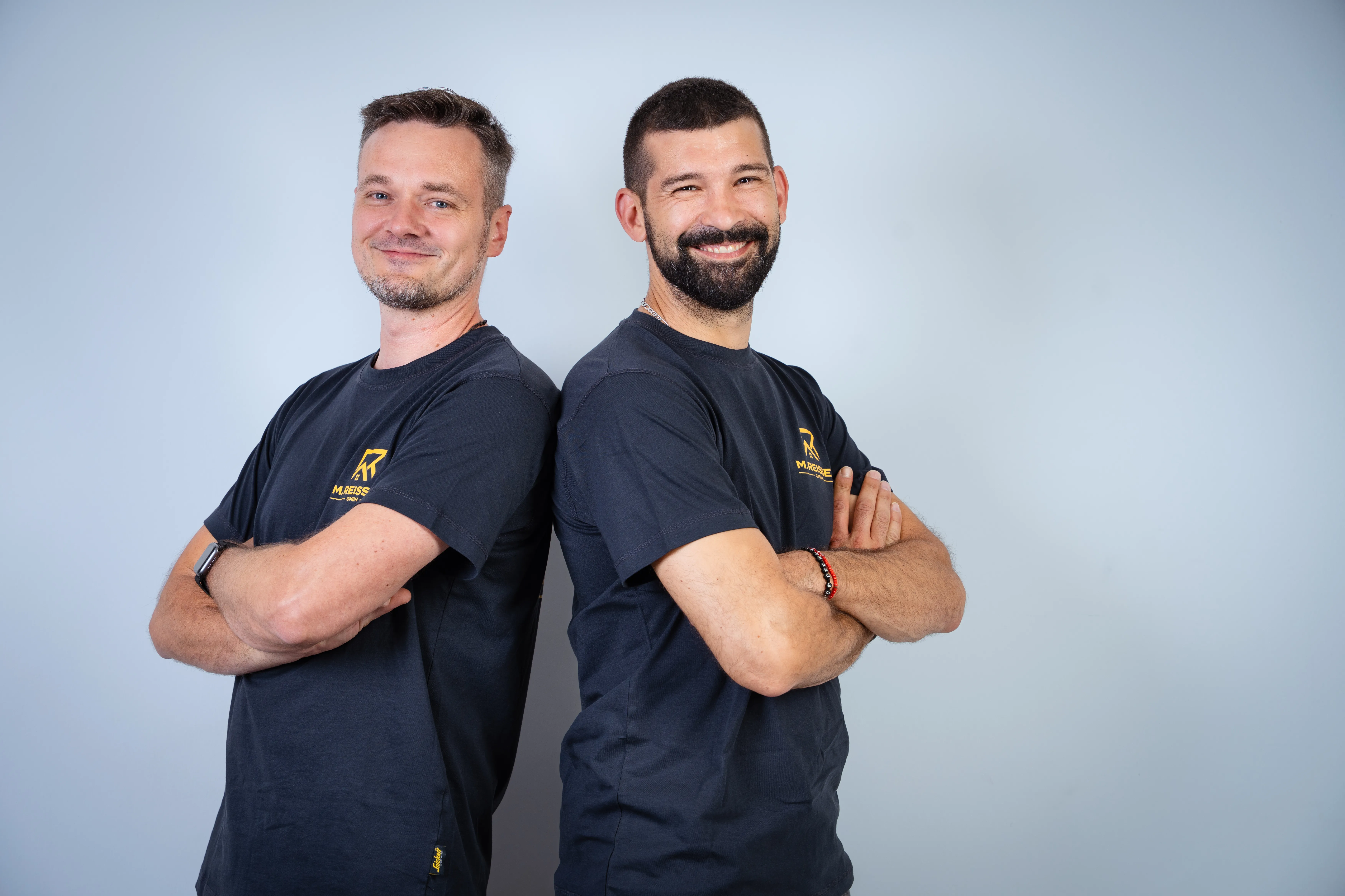 Zwei Männer stehen Rücken an Rücken mit verschränkten Armen und lächeln in schwarzen Shirts.