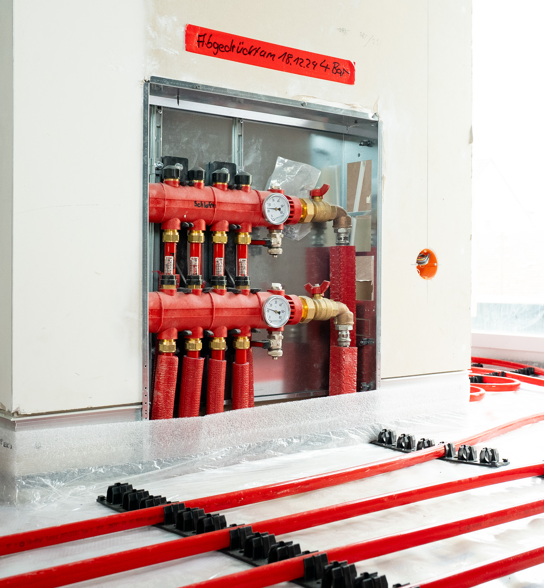 Installation einer roten Flächenheizung mit Rohren und zwei Druckmessgeräten in einer Wandöffnung.