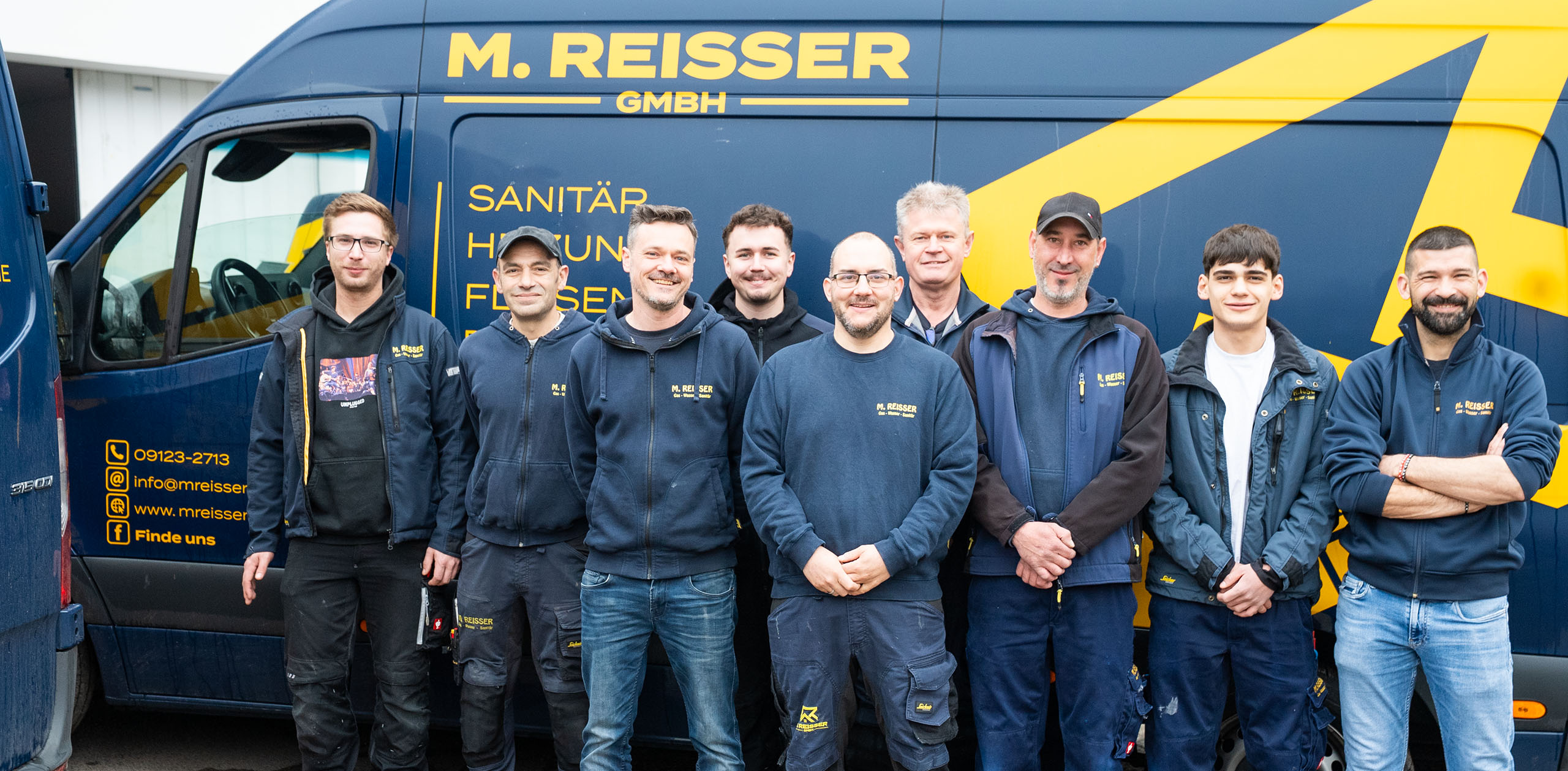 Acht Männer in Arbeitskleidung stehen vor einem blauen Firmenfahrzeug von M. Reisser GmbH.