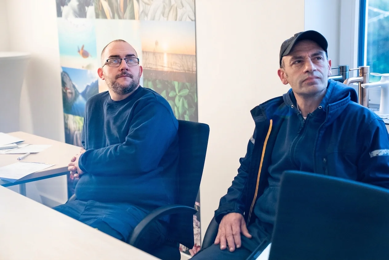 Zwei Männer sitzen an einem Tisch in einem Raum mit Fotos an der Wand und schauen in verschiedene Richtungen.