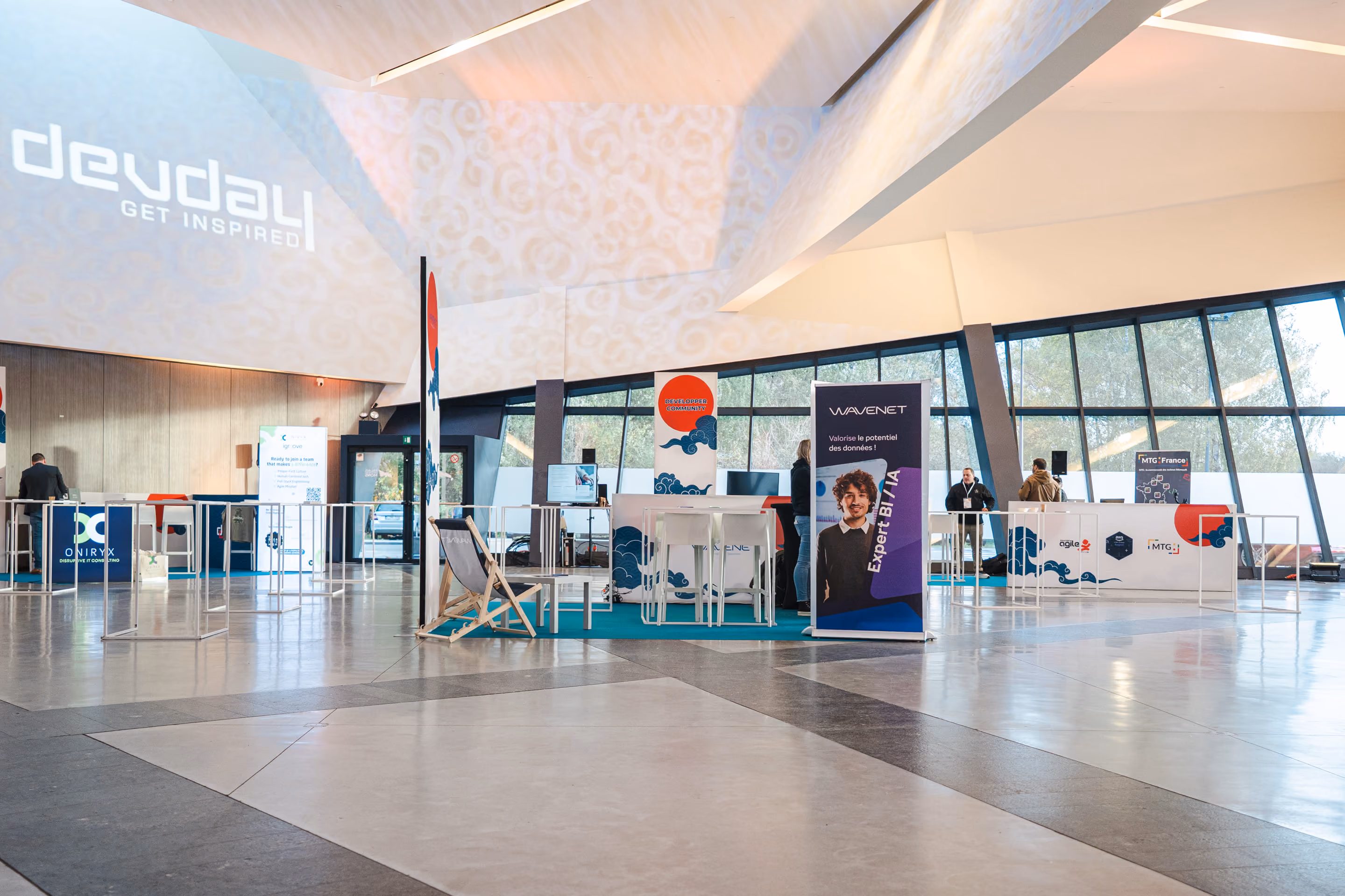 Espace d'exposition lumineux avec plusieurs stands et panneaux publicitaires, dont un pour WAVENET vantant le potentiel des données et un autre pour devdai avec le slogan "GET INSPIRED" projeté au mur.