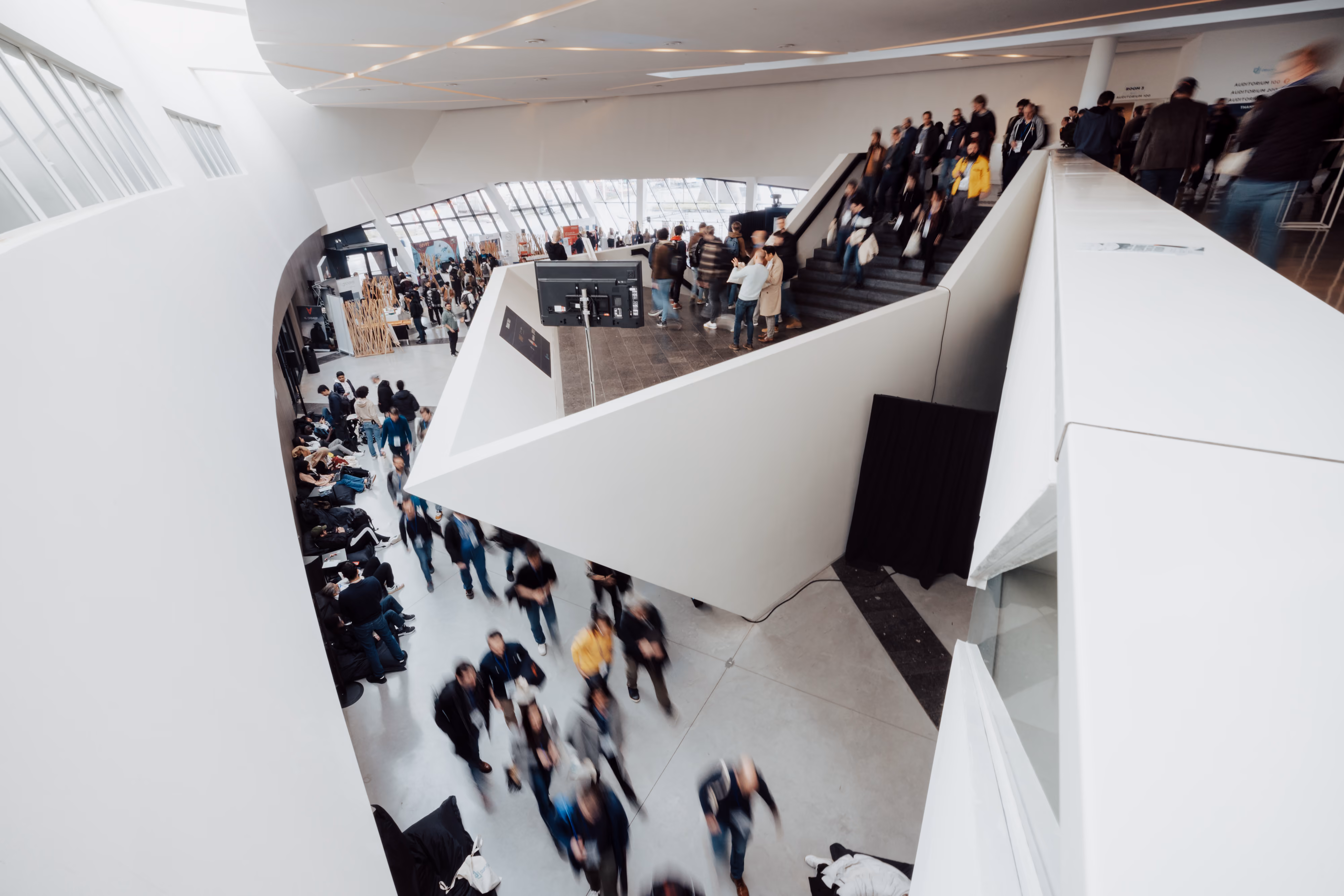 Un espace intérieur moderne et spacieux avec beaucoup de lumière naturelle où se déplacent de nombreuses personnes, certaines montant et descendant un escalier central blanc à la structure géométrique marquée.