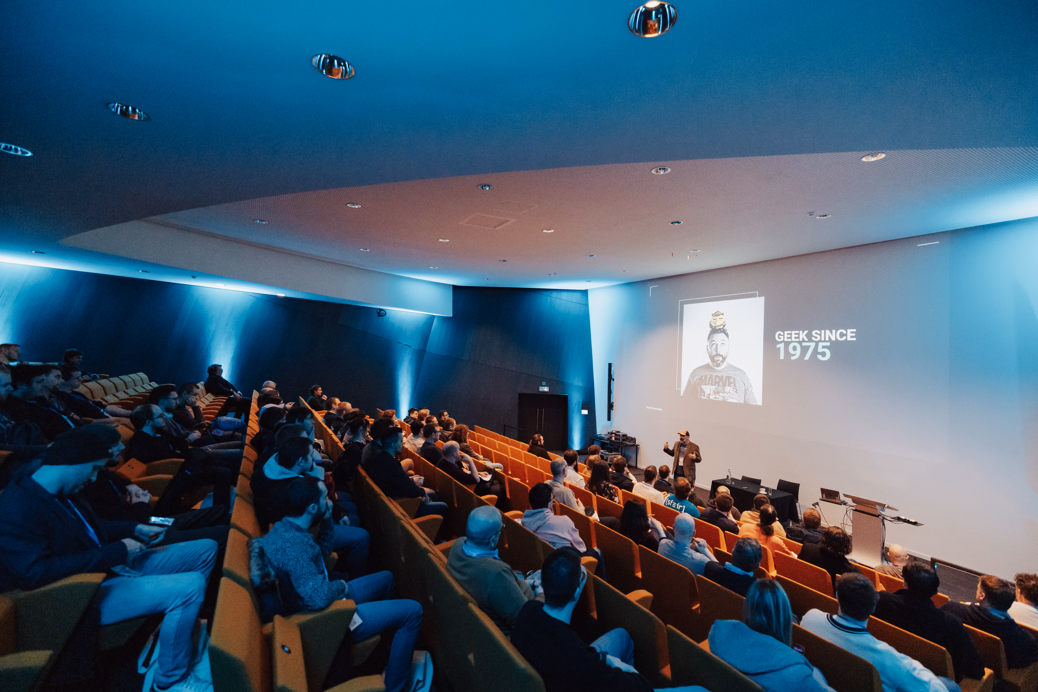 Une salle de conférence sombre avec plusieurs rangées de sièges orange occupés par un public écoutant un orateur debout devant un mur blanc où est projetée une image avec le texte 'GEEK SINCE 1975'.