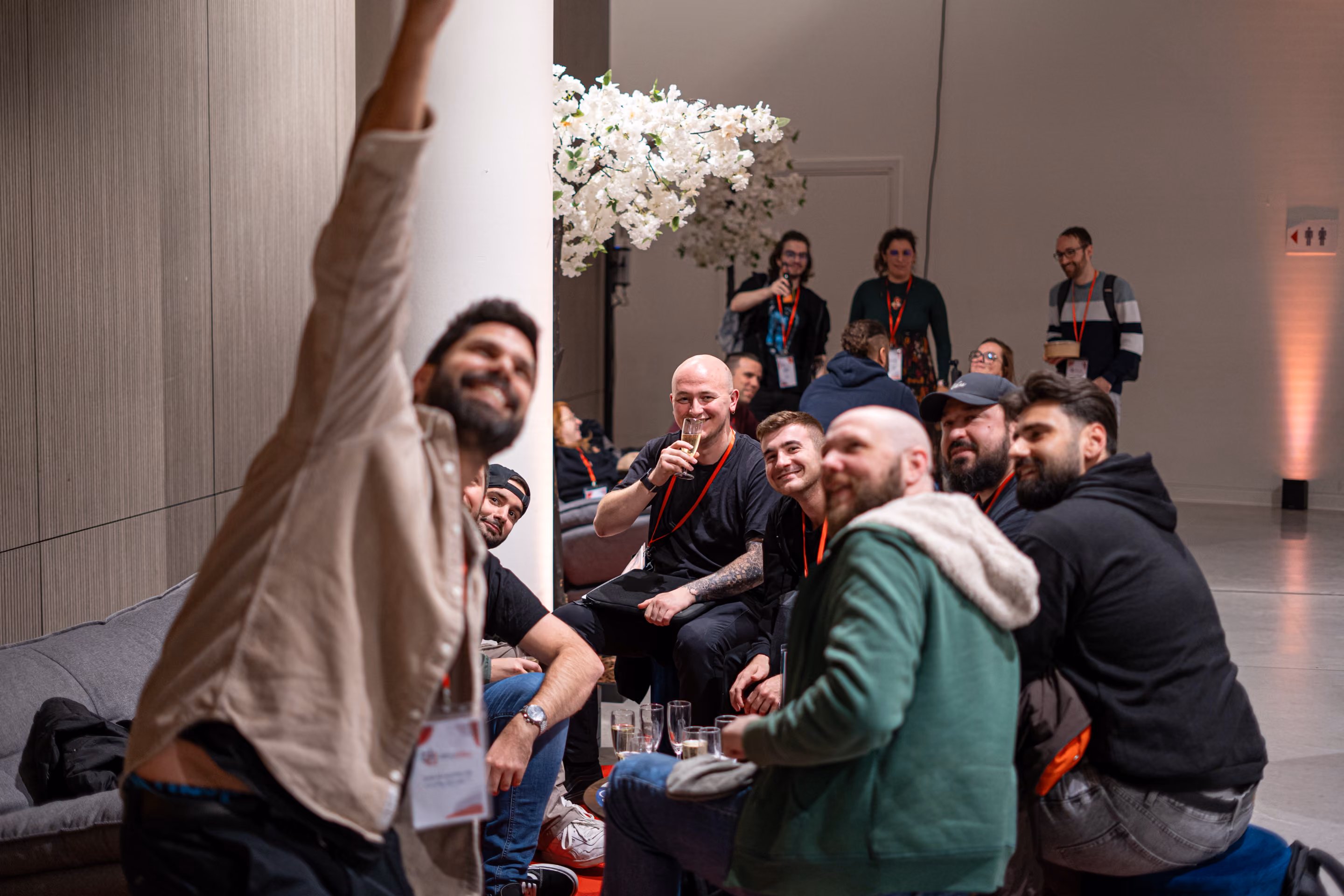Un groupe d'hommes assis sur des canapés et des coussins dans un espace intérieur moderne, certains tenant des verres à vin ou à champagne, posant pour un selfie pris par un homme au premier plan portant un badge autour du cou, avec des décorations florales blanches en arrière-plan et d'autres personnes debout à l'arrière-plan.