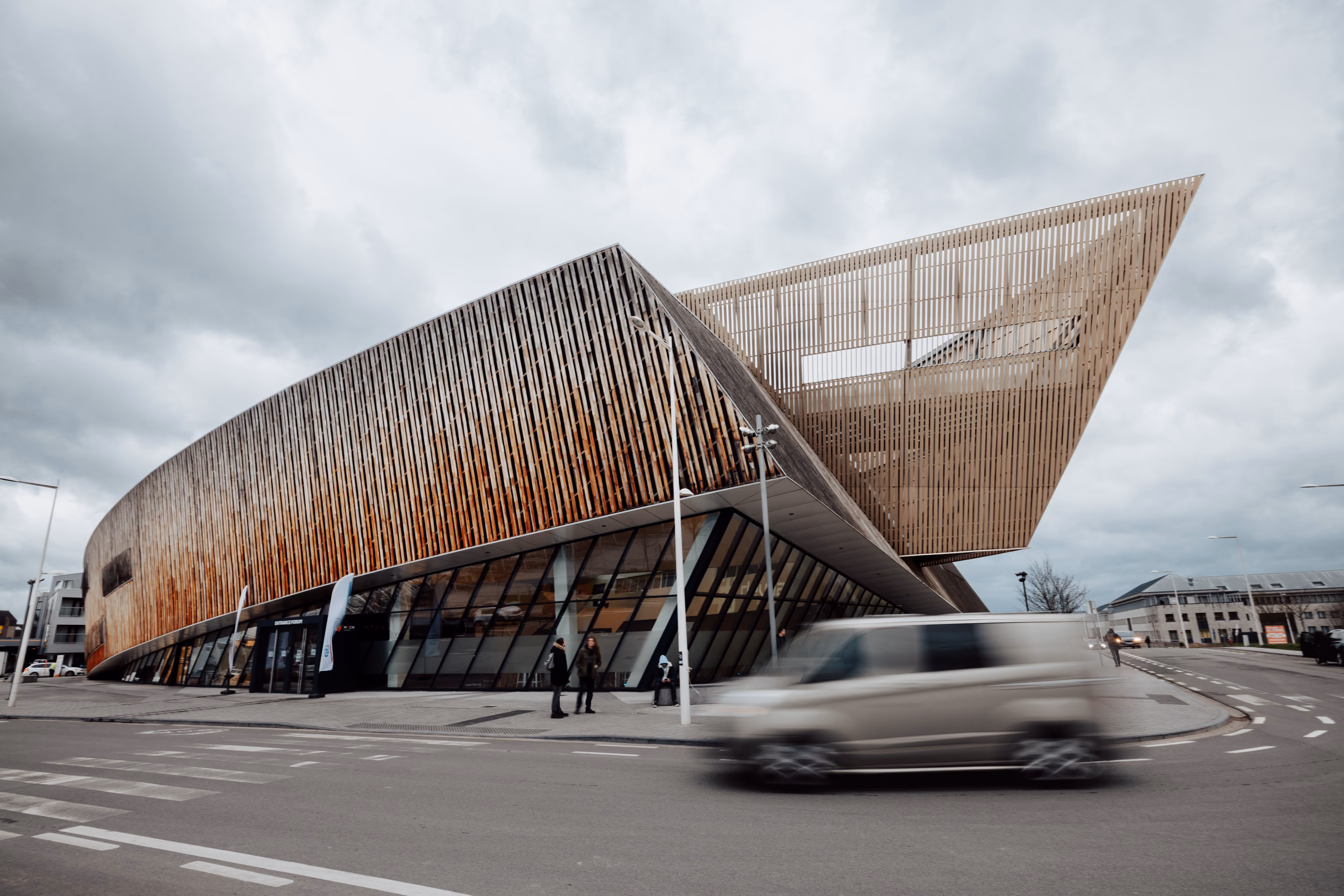 Bâtiment moderne en bois avec une grande structure angulaire en vol, situé à un coin de rue avec une voiture grise en mouvement flou au premier plan et des passants devant l'entrée.