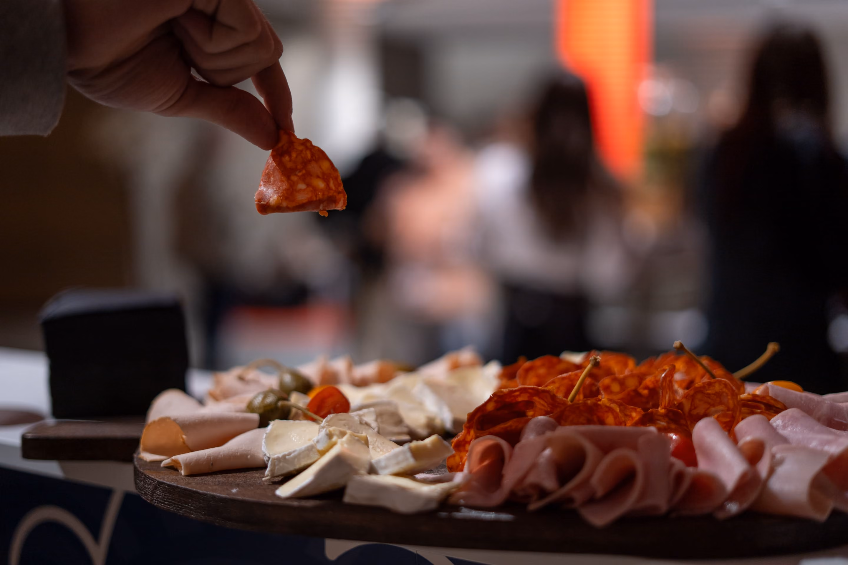 Une main tient une tranche de charcuterie épicée au-dessus d'un plateau en bois avec du fromage, des olives et différentes charcuteries, en arrière-plan flou de personnes.