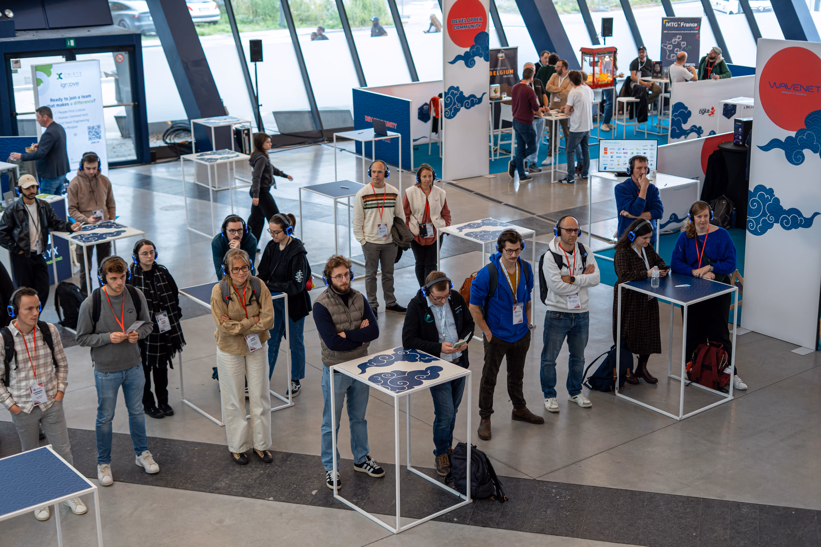 Groupe de personnes portant des écouteurs et badge lors d'une conférence ou exposition en intérieur, certaines regardent vers l'avant, d'autres utilisent un téléphone, avec des stands et panneaux de sponsors en arrière-plan.