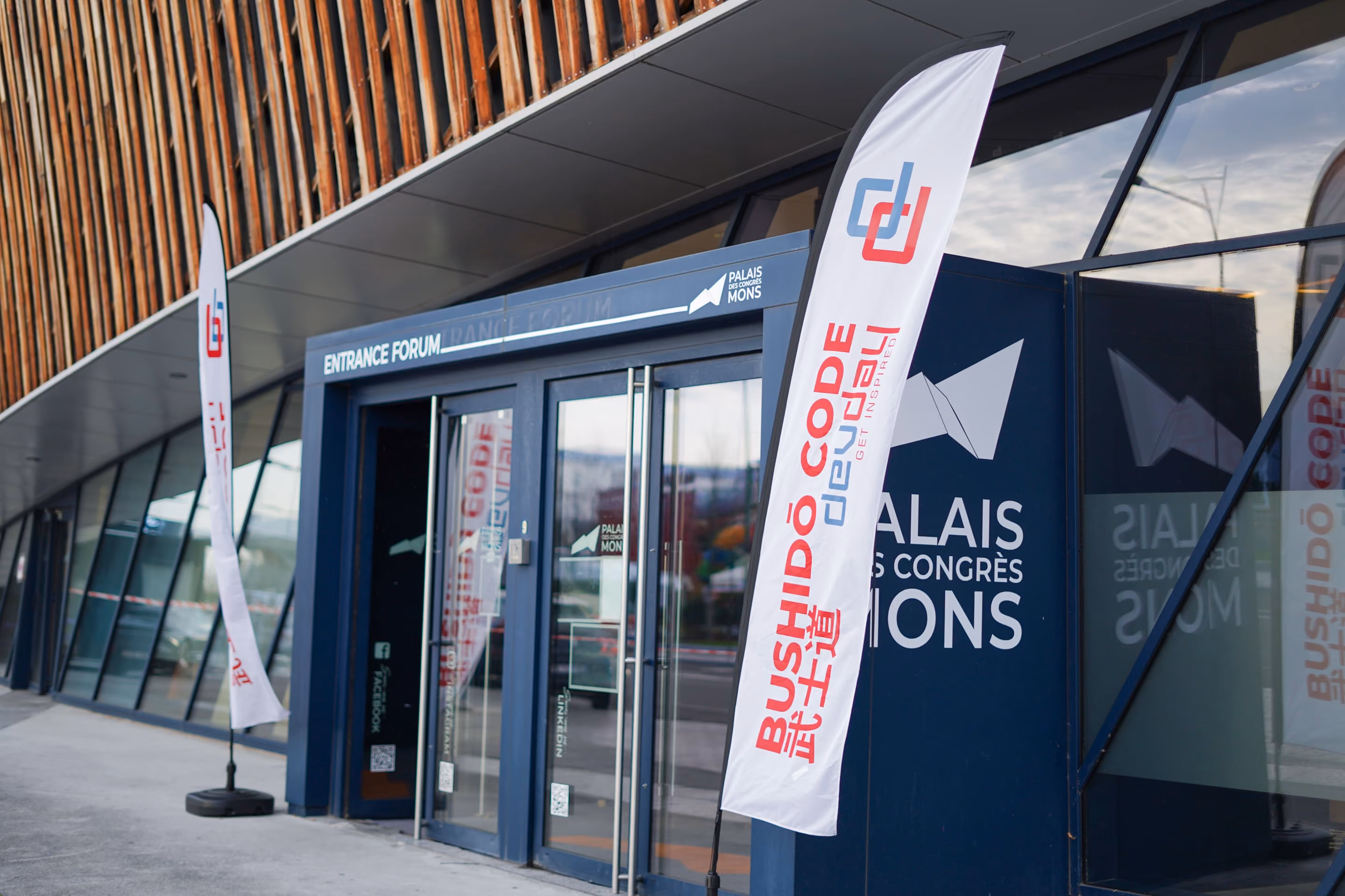 Entrée du Palais des Congrès Mons avec deux drapeaux blancs portant l'inscription Bushidô Code Duel Get Inspired en rouge et bleu, devant une façade moderne en bois et verre.