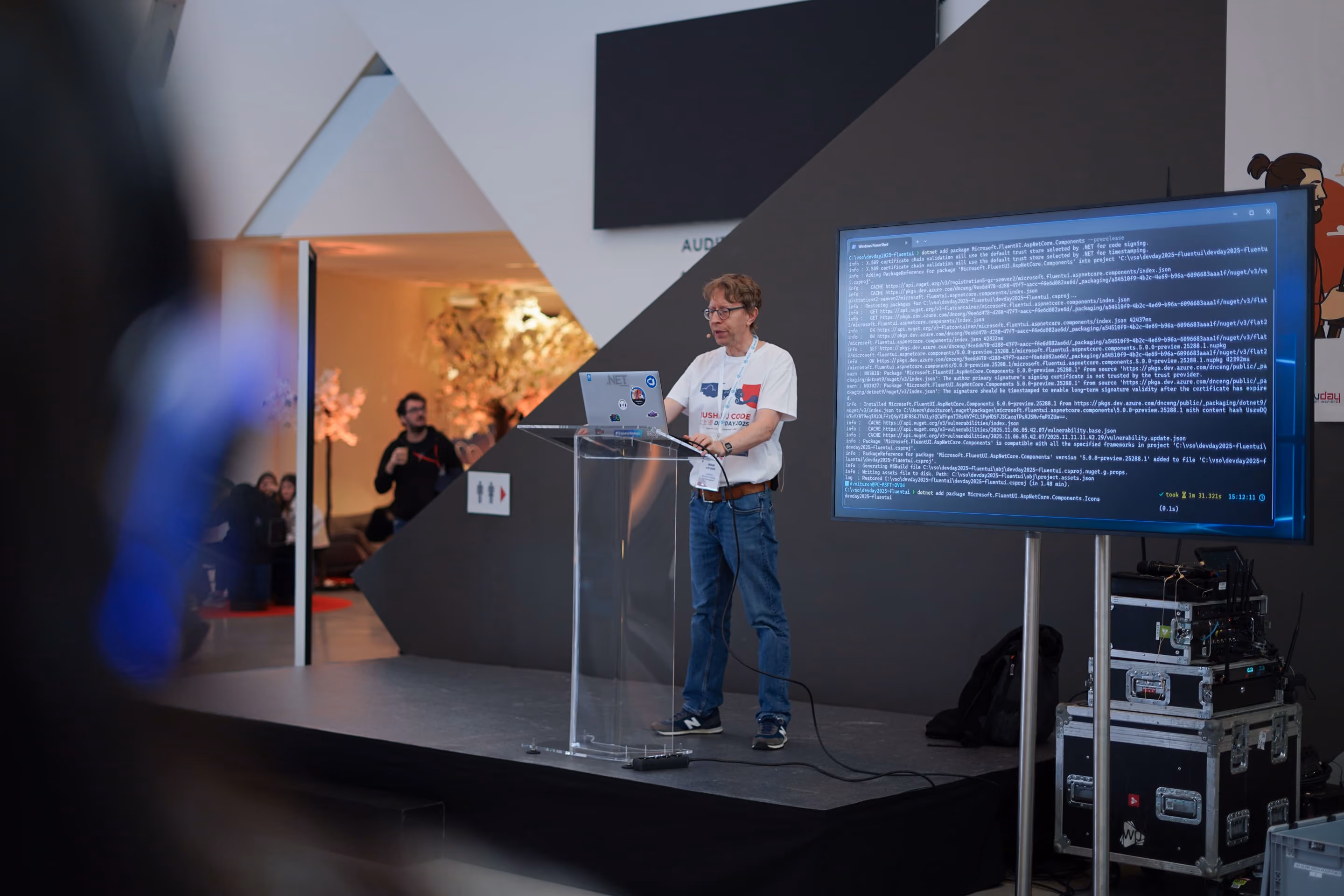 Un homme porte un t-shirt blanc avec un badge de conférence et fait une présentation sur scène avec un ordinateur portable sur un pupitre transparent, un grand écran affichant du code, et un public en arrière-plan dans un espace moderne.