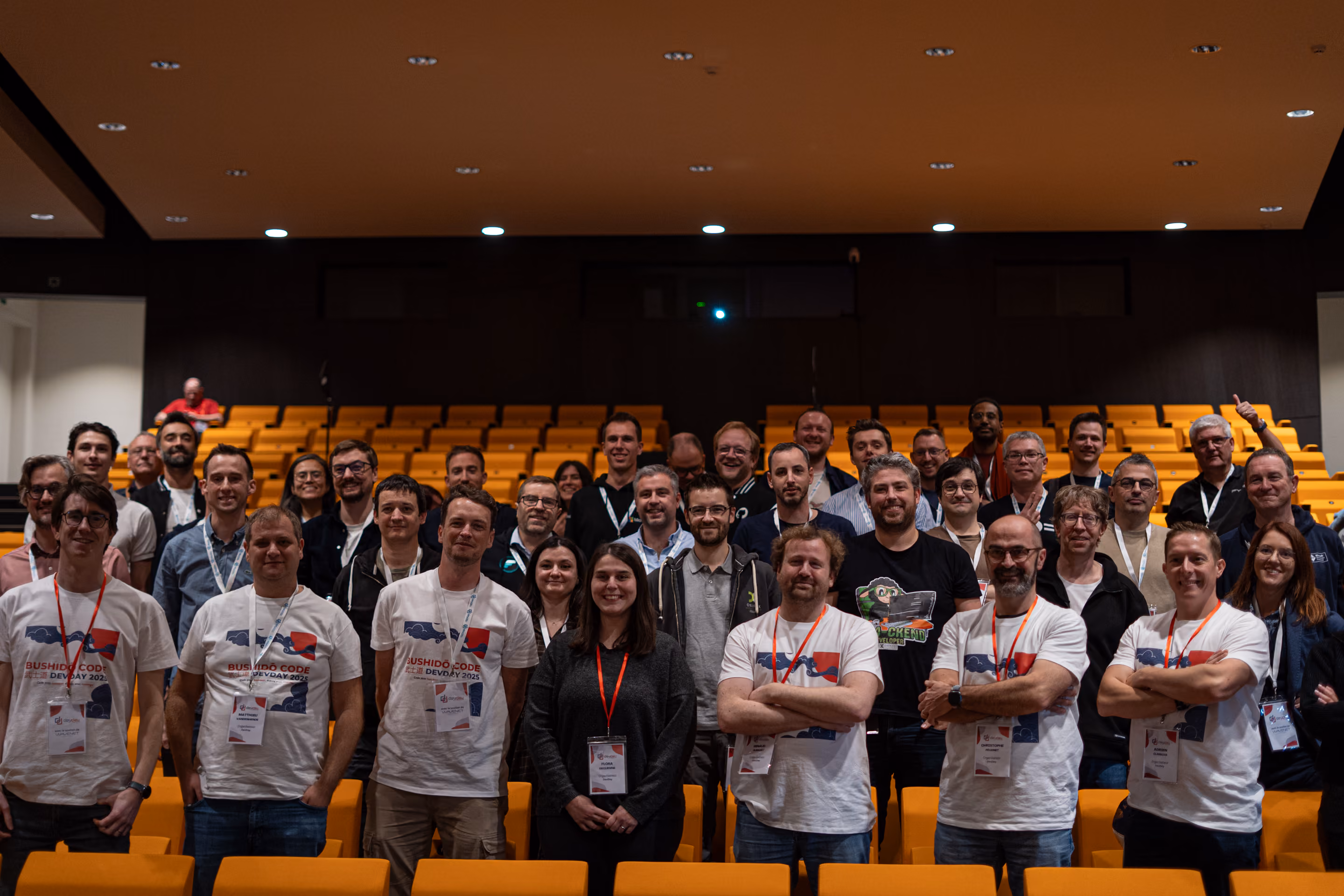 Groupe de personnes posant dans une salle de conférence avec des sièges jaunes, plusieurs portant des t-shirts blancs avec le logo Bushido Code DevDay 2023 et des badges d'identification.