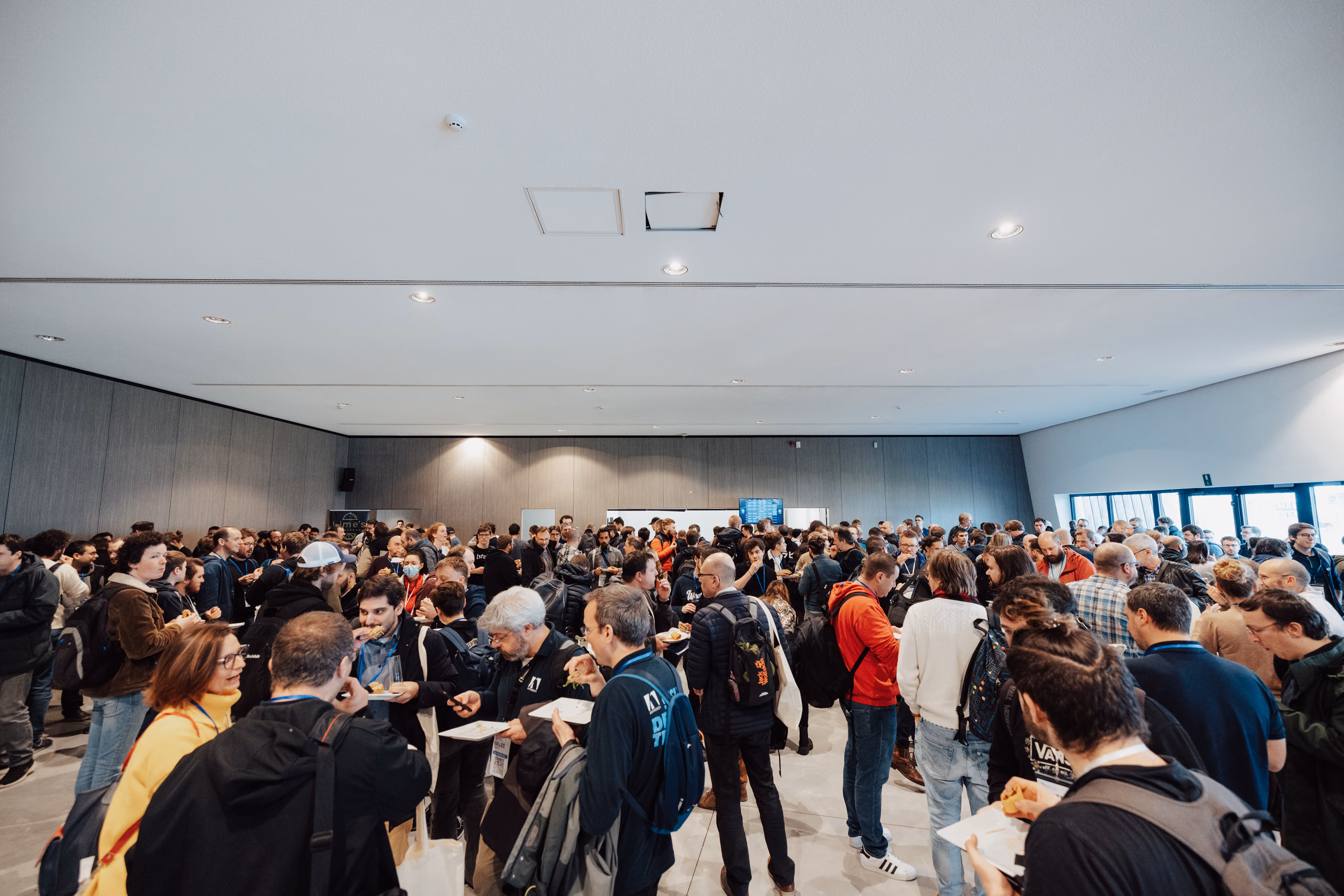 Grande foule de personnes adultes dans une salle moderne, certaines portent des badges et tiennent des assiettes avec de la nourriture, discutant et socialisant sous un plafond blanc avec éclairage encastré.