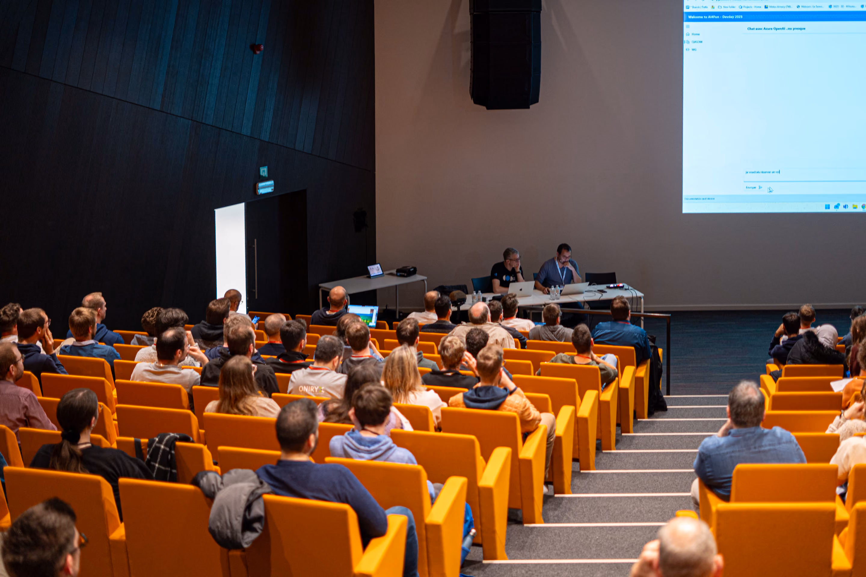 Une salle de conférence avec des rangées de sièges orange occupés par des participants regardant deux intervenants assis à une table avec des ordinateurs portables devant un écran projeté affichant une interface de chat en ligne.