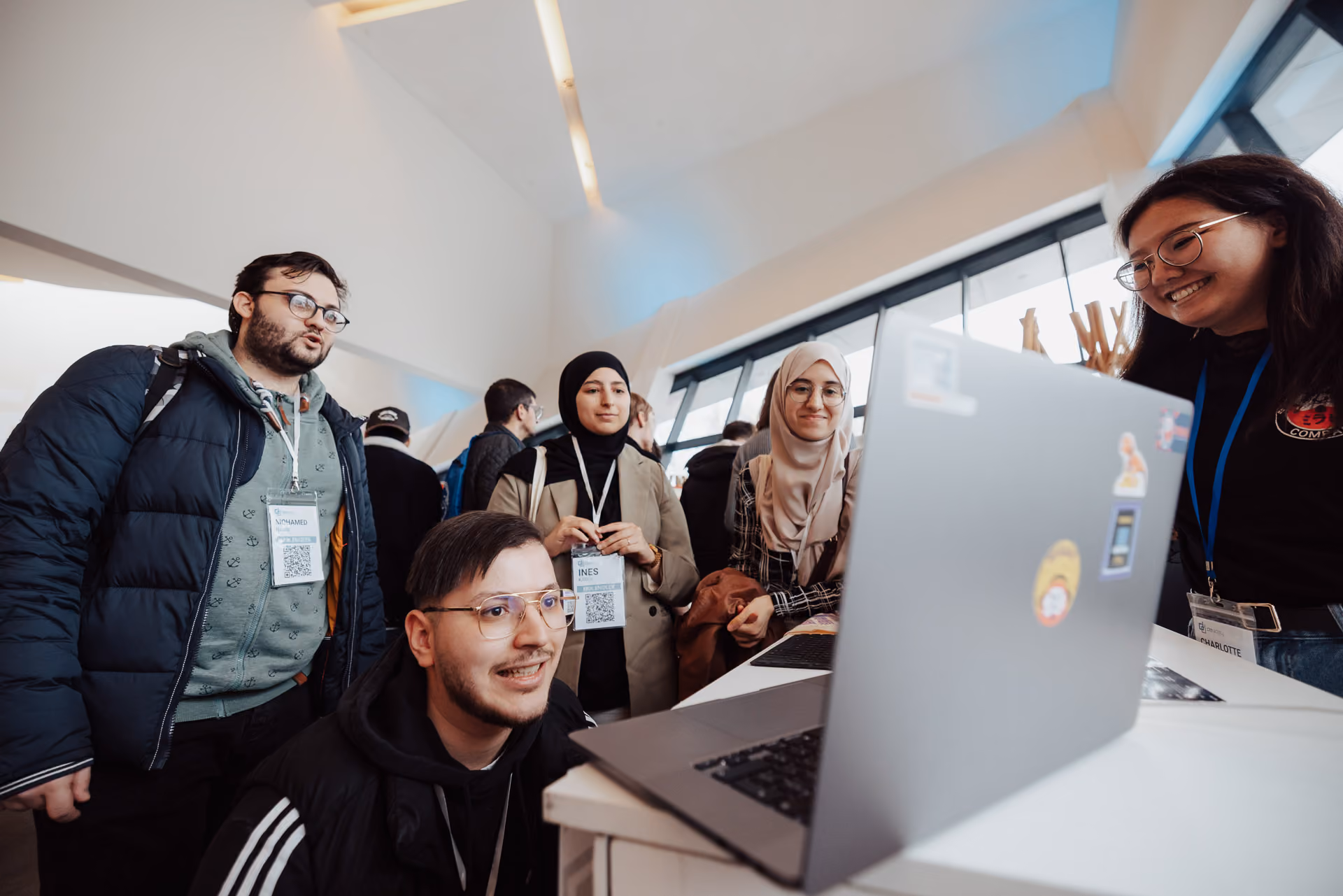 Groupe de cinq jeunes personnes souriantes et attentives, regardant un ordinateur portable sur une table dans un espace intérieur moderne, avec de grandes fenêtres en arrière-plan.